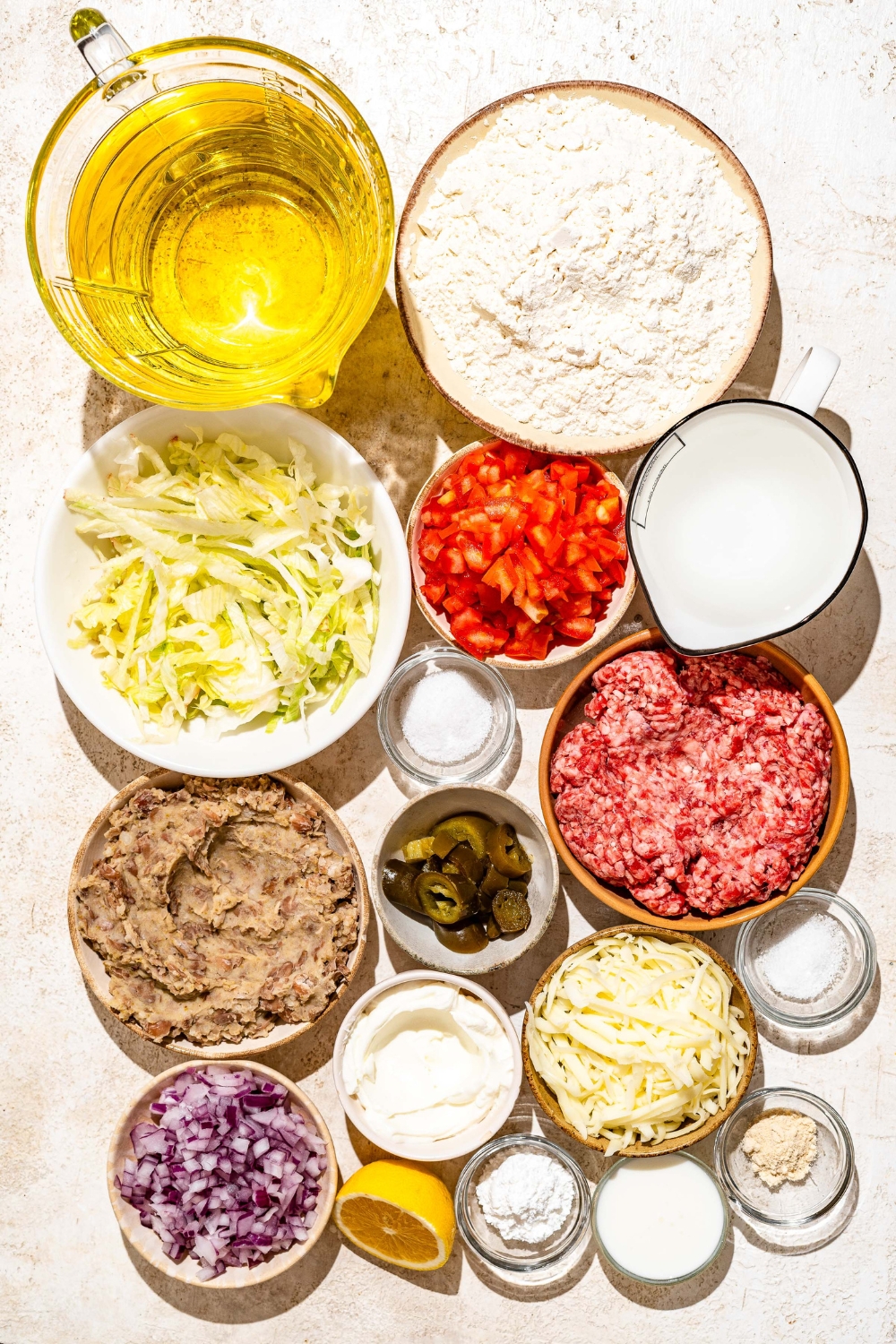 A tan counter with several bowls containing ingredients to make Navajo tacos including oil, flour, baking powder, salt, tomatoes, ground beef, pinto beans, mayo, lemon, onion, and more.