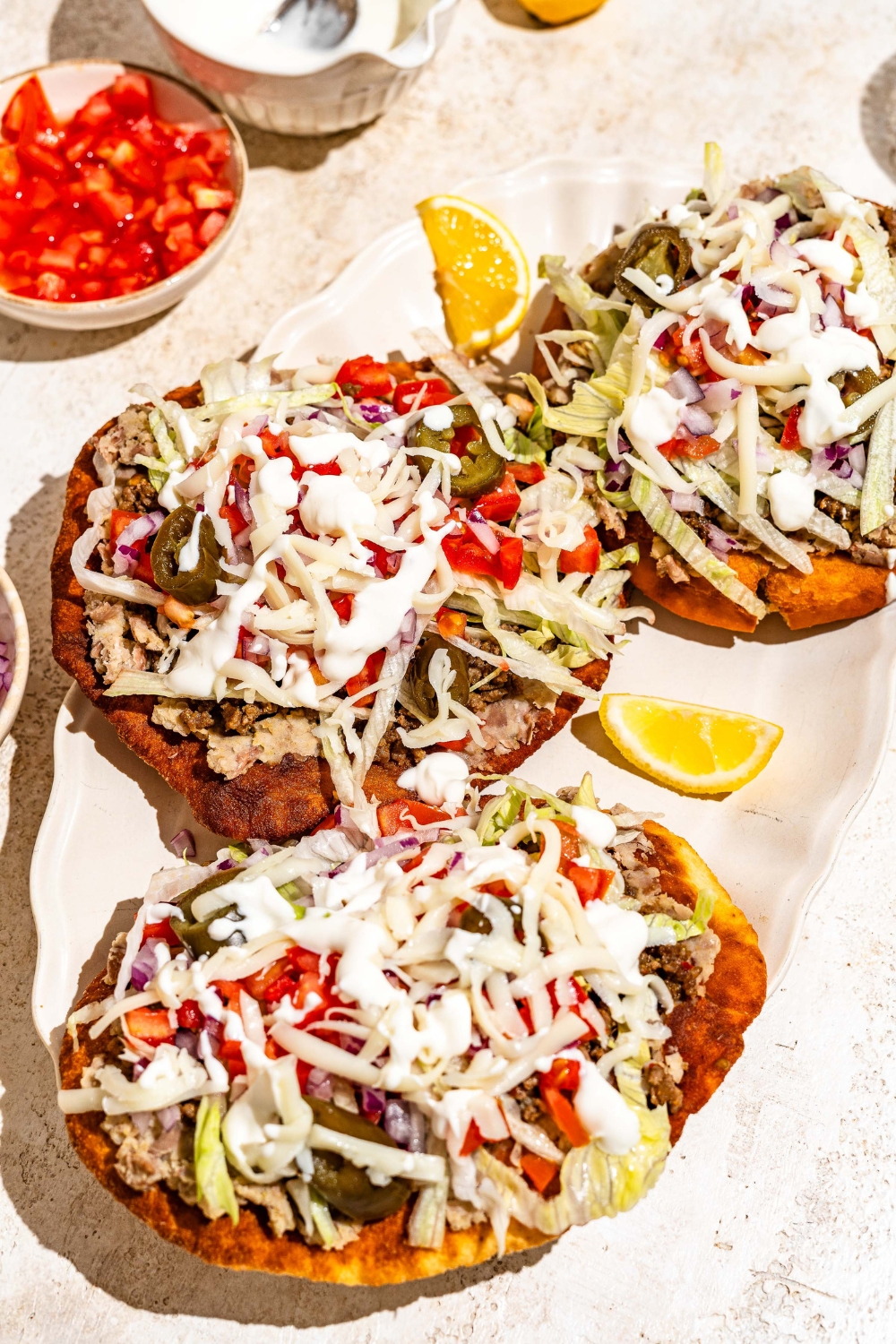 A white platter with several Navajo tacos with Indian fry bread, ground beef, beans, jalapeños, and shredded cabbage with white sauce drizzled on top. The platter is served with sliced lemon. The platter is on a tan counter with bowls of taco toppings.