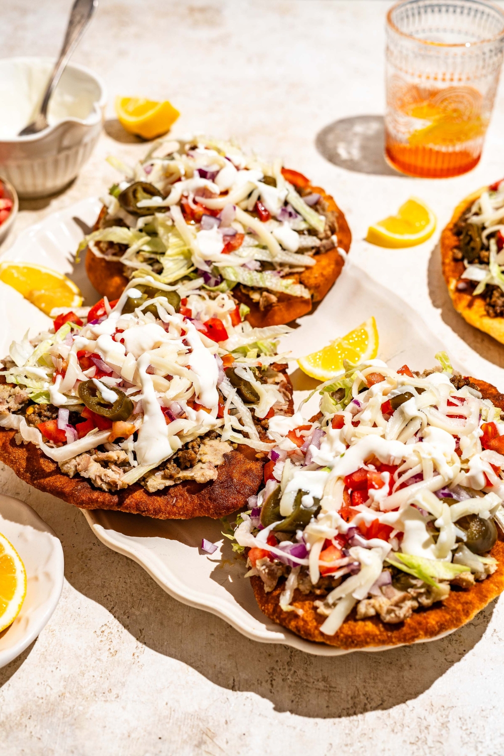 A white platter with several Navajo tacos with Indian fry bread, ground beef, beans, jalapeños, and shredded cabbage with white sauce drizzled on top. The platter is served with sliced lemon.