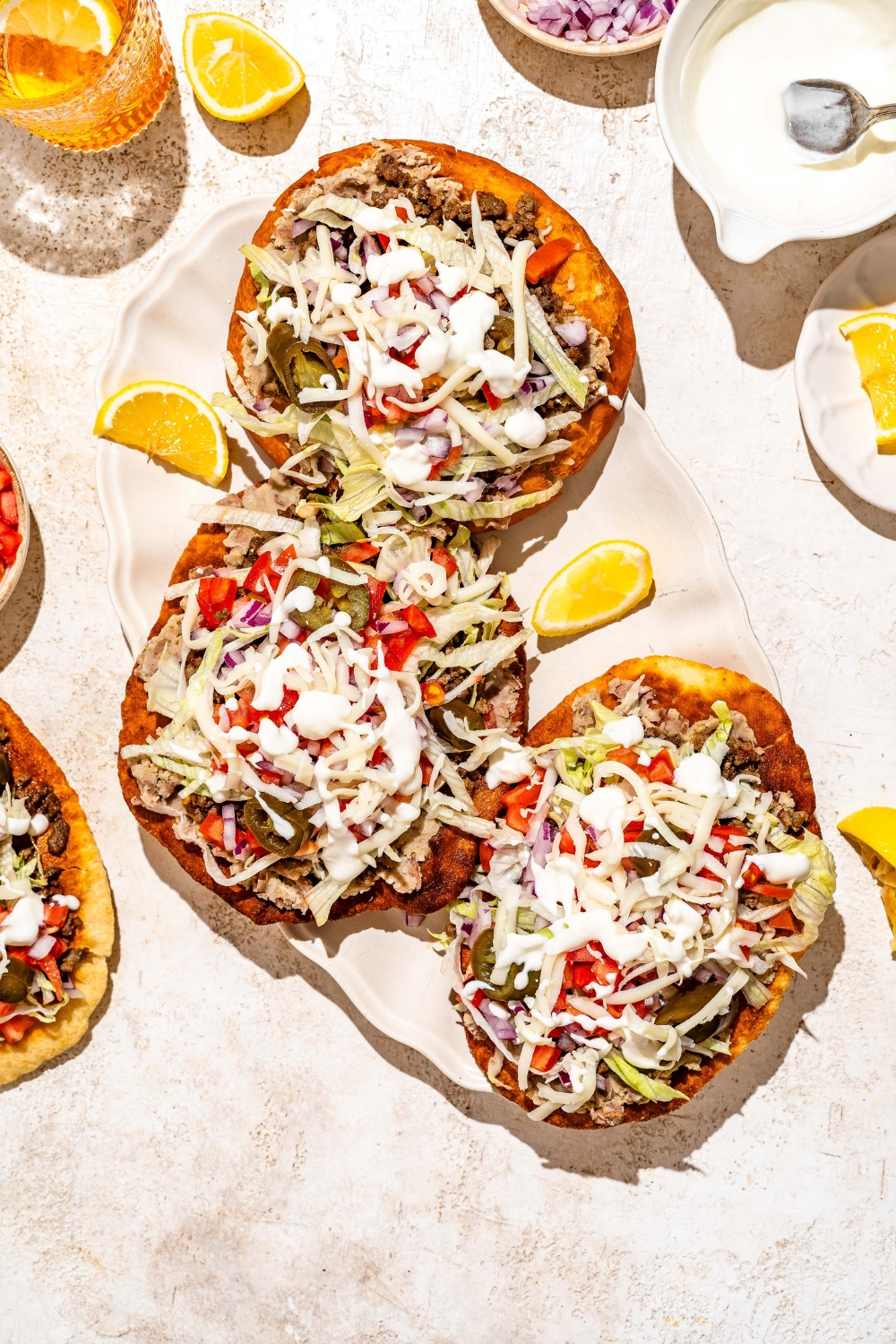 A white platter with several Navajo tacos with Indian fry bread, ground beef, beans, jalapeños, and shredded cabbage with white sauce drizzled on top. The platter is served with sliced lemon. The platter is on a tan counter with bowls of taco toppings.