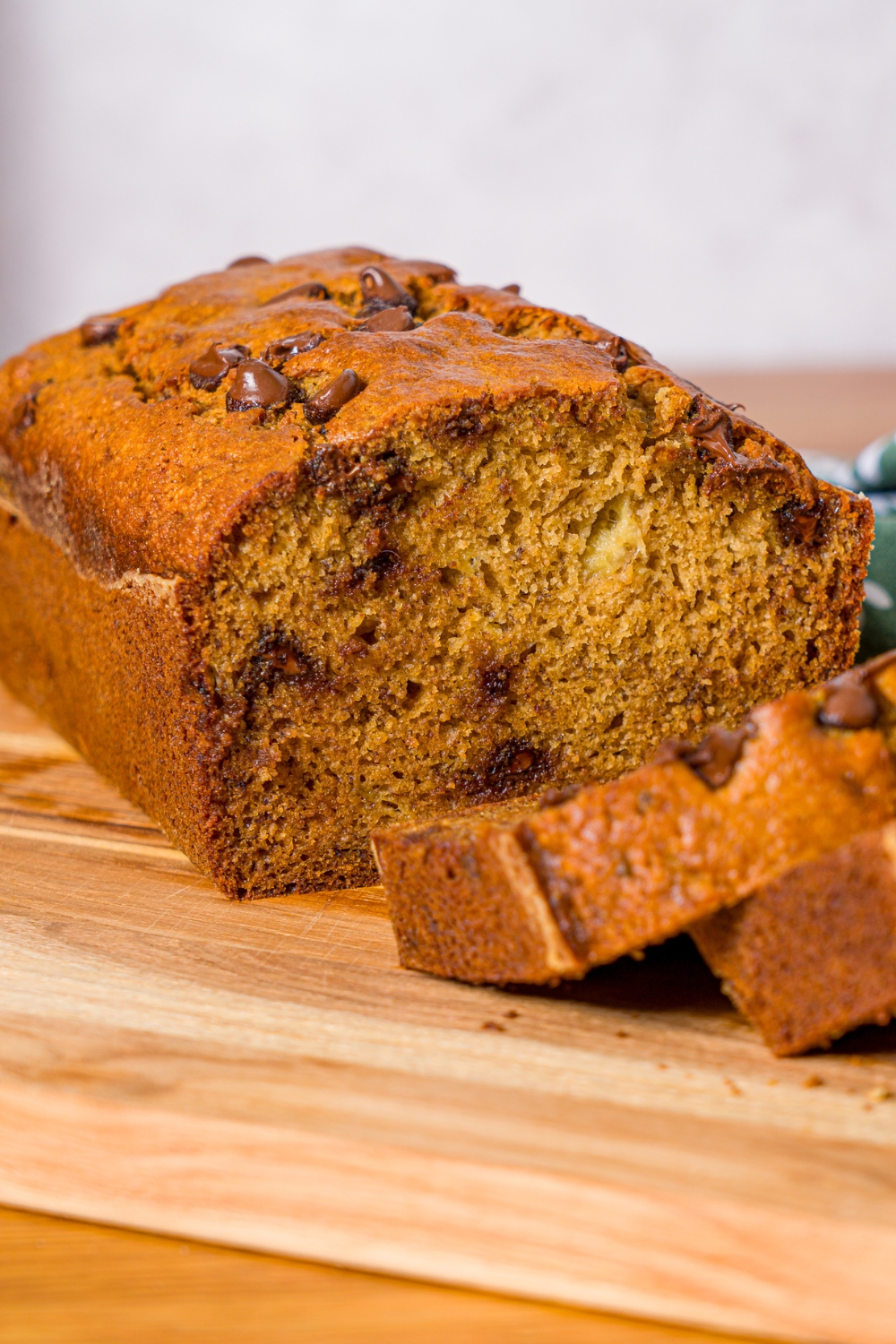A wooden board with a loaf of chocolate chip banana bread with olive oil. Several slices have been sliced from the loaf. The board is on a wooden counter with a green floral napkin.