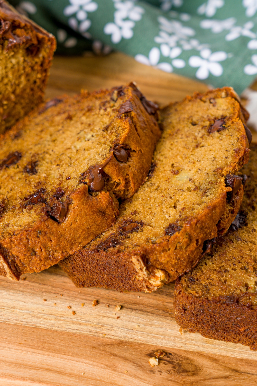 A wooden board with a loaf of chocolate chip banana bread with olive oil. Several slices have been sliced from the loaf. The board is on a wooden counter with a green floral napkin.