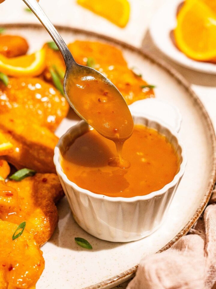 A white plate with chicken tossed in orange sauce with a small bowl of orange sauce. A spoon is dipping in the bowl. The plate is on a tan counter with a brown cloth napkin.