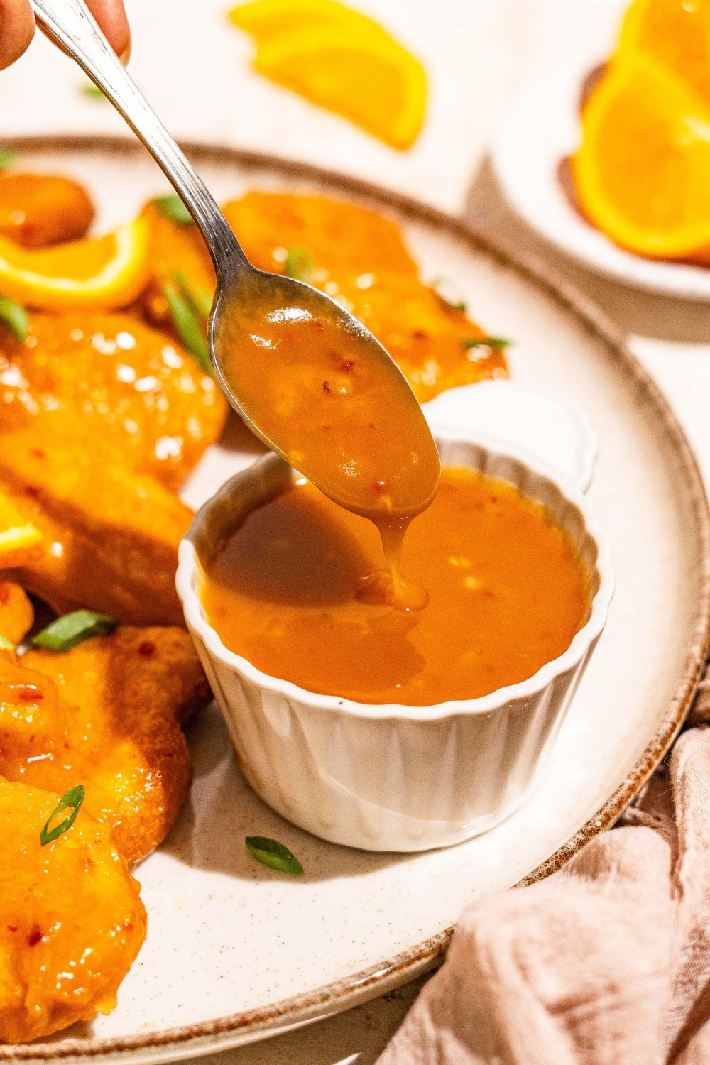 A white plate with chicken tossed in orange sauce with a small bowl of orange sauce. A spoon is dipping in the bowl. The plate is on a tan counter with a brown cloth napkin.