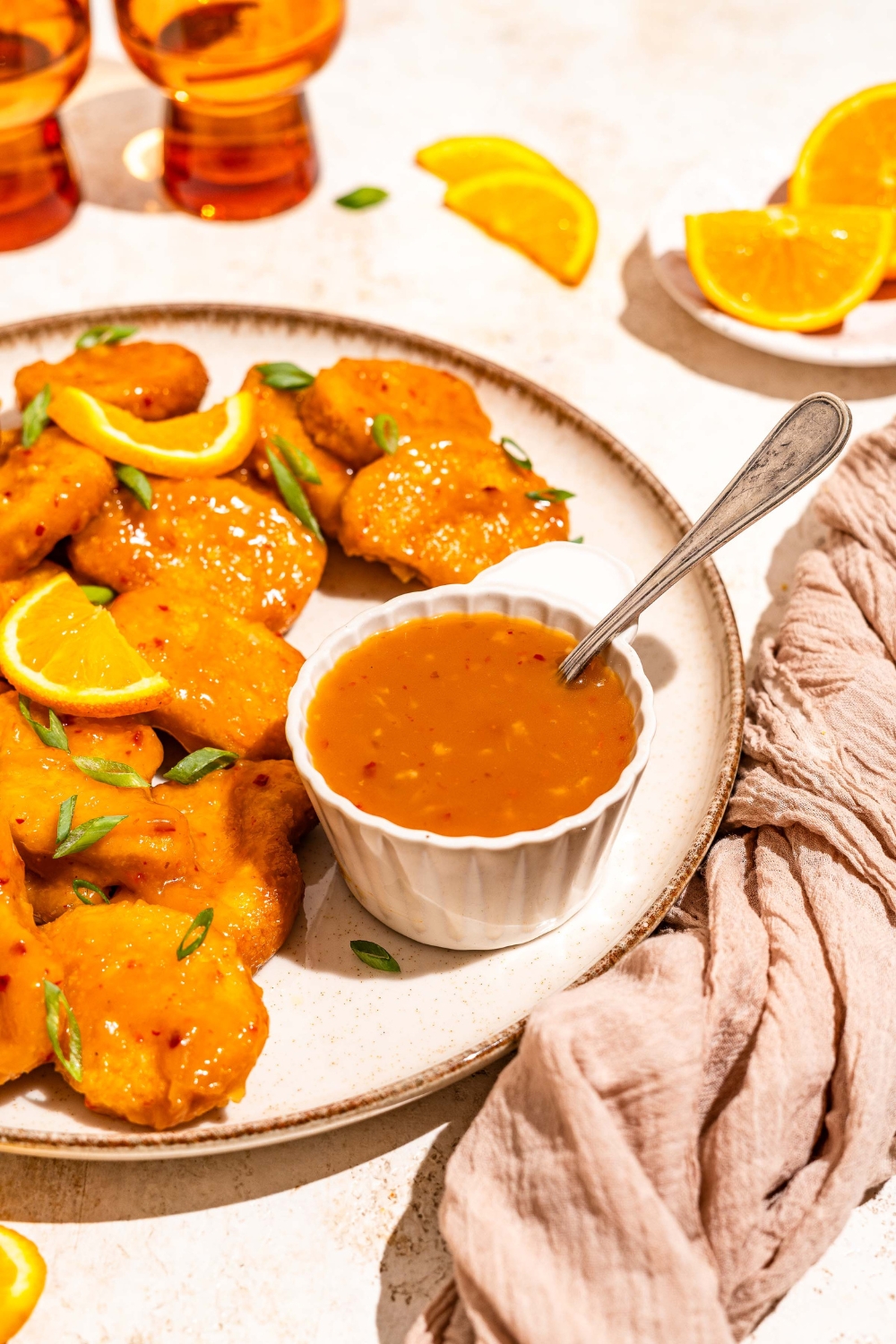 A white plate with chicken tossed in orange sauce and garnished with sliced green onion and orange slices. The plate is served with a small bowl of orange sauce with a spoon in the bowl. The plate is on a tan counter with a brown cloth napkin.