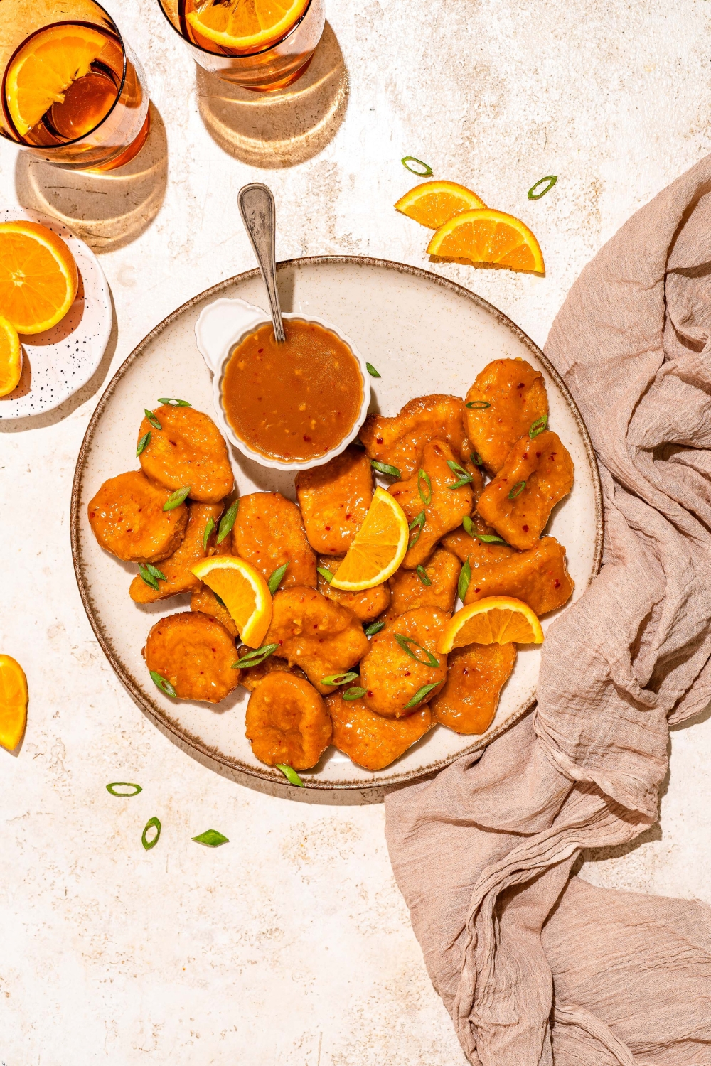A white plate with chicken tossed in orange sauce and garnished with sliced green onion and orange slices. The plate is served with a small bowl of orange sauce with a spoon in the bowl. The plate is on a tan counter with a brown cloth napkin.