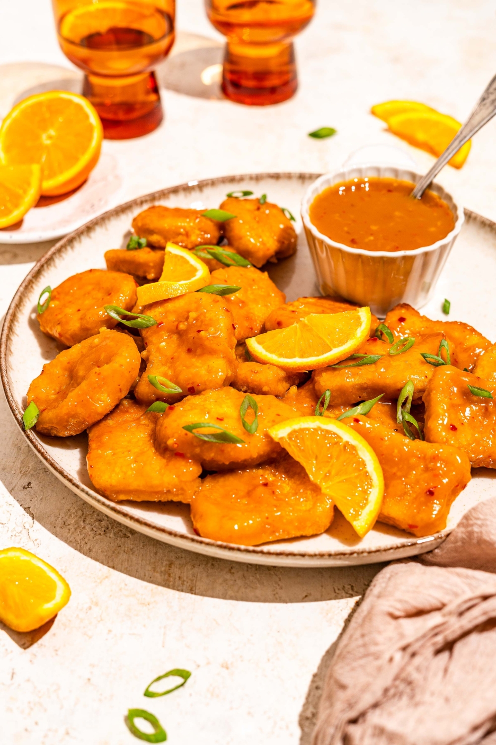 A white plate with chicken tossed in orange sauce and garnished with sliced green onion and orange slices. The plate is served with a small bowl of orange sauce with a spoon in the bowl. The plate is on a tan counter with a brown cloth napkin.