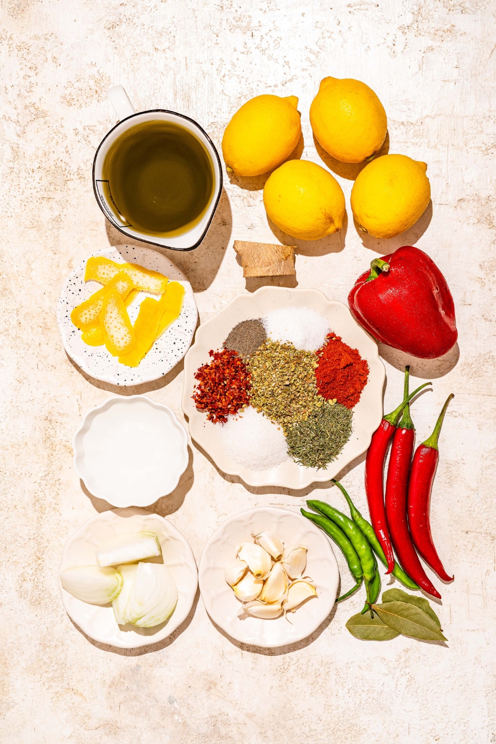 A tan counter with several bowls containing ingredients to make peri peri sauce including lemon, bell pepper, chilies, garlic, onion, sugar, and spices.