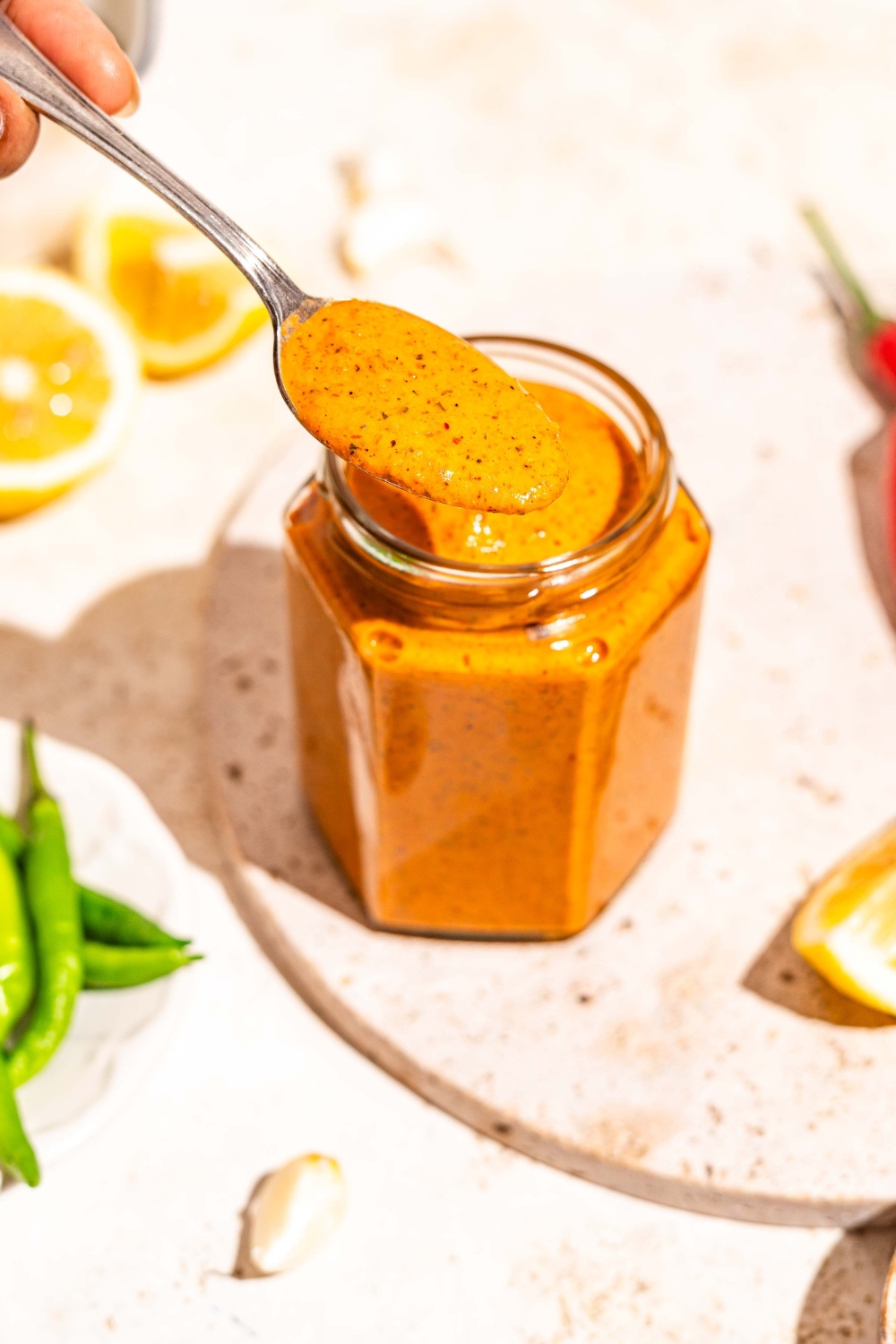 A jar of peri peri sauce on a marble slab. A spoon is dipping into the jar. The slab is on a tan counter with sliced lemon.