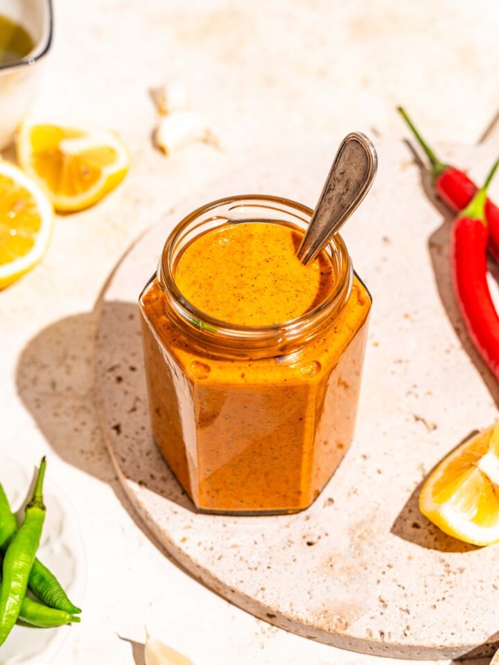 A jar of peri peri sauce on a marble slab with a spoon in the jar. The slab is on a tan counter with sliced lemon and chilies.