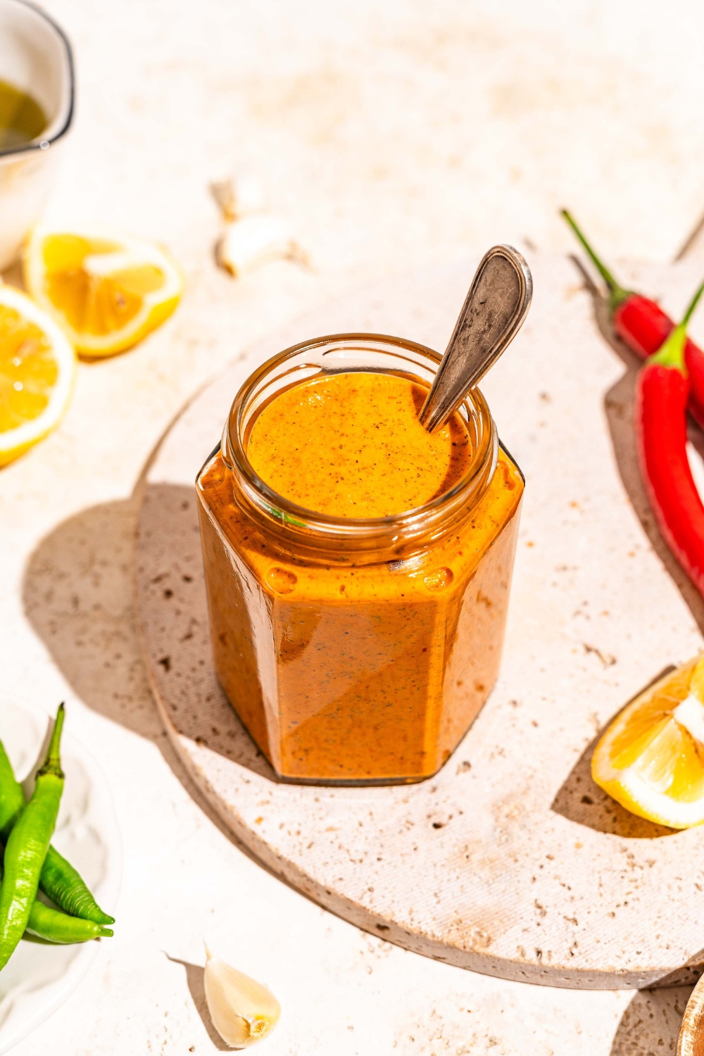 A jar of peri peri sauce on a marble slab with a spoon in the jar. The slab is on a tan counter with sliced lemon and chilies.
