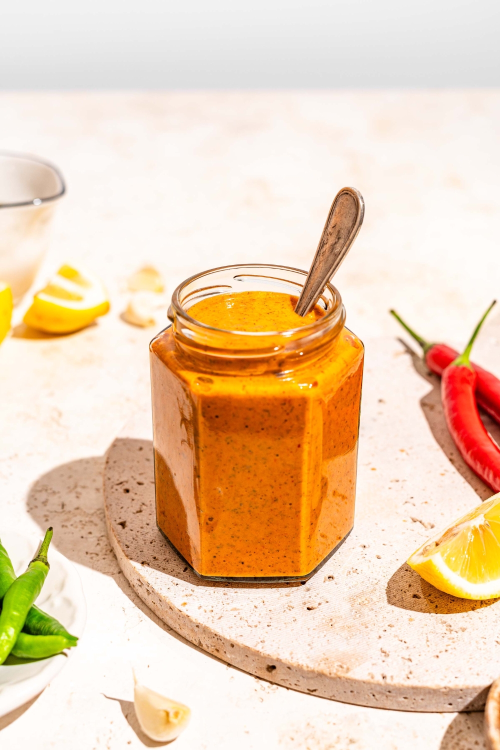 A jar of peri peri sauce on a marble slab with a spoon in the jar. The slab is on a tan counter with sliced lemon and chilies.