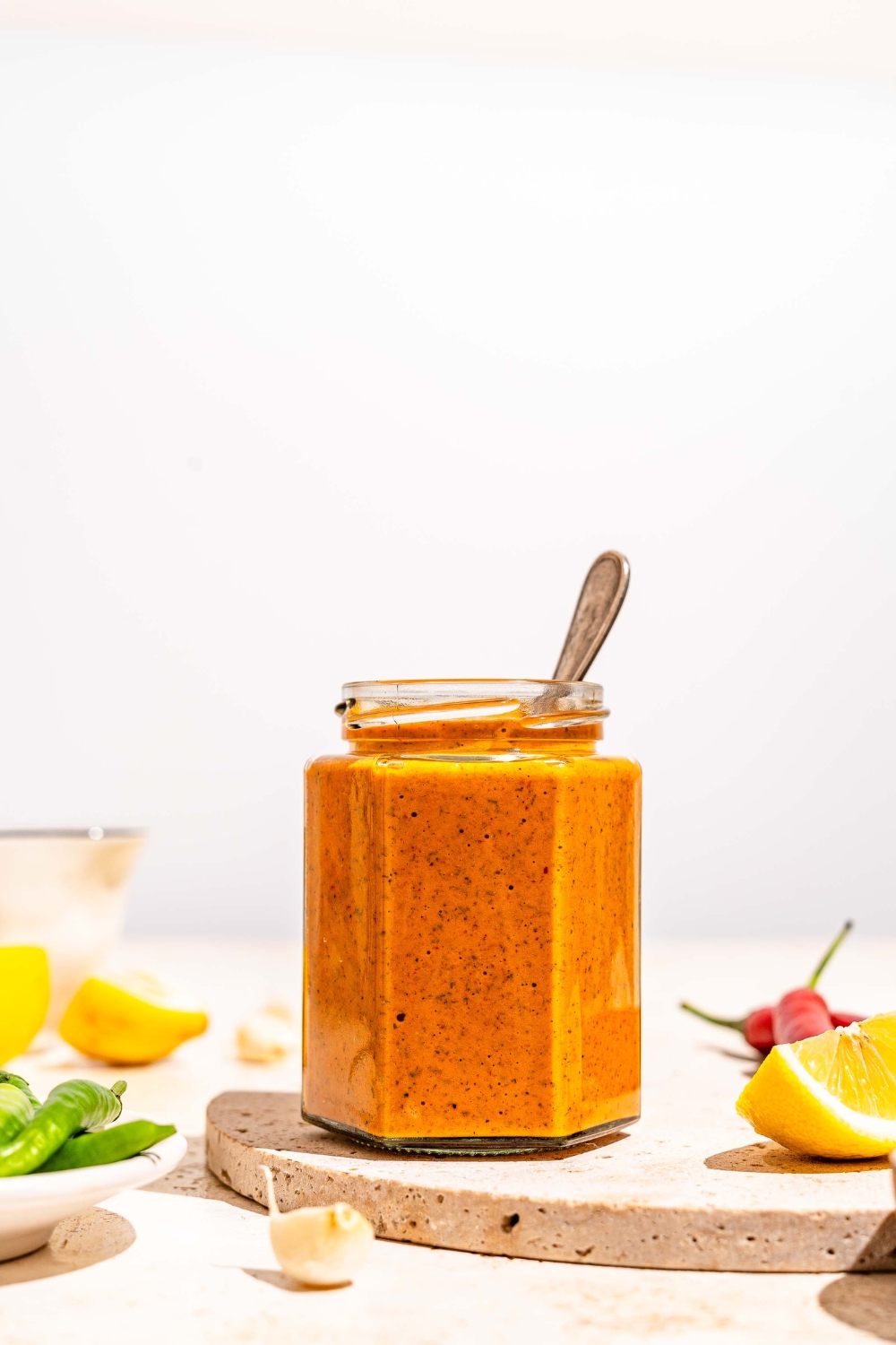 A jar of peri peri sauce on a marble slab with a spoon in the jar. The slab is on a tan counter with sliced lemon and chilies.