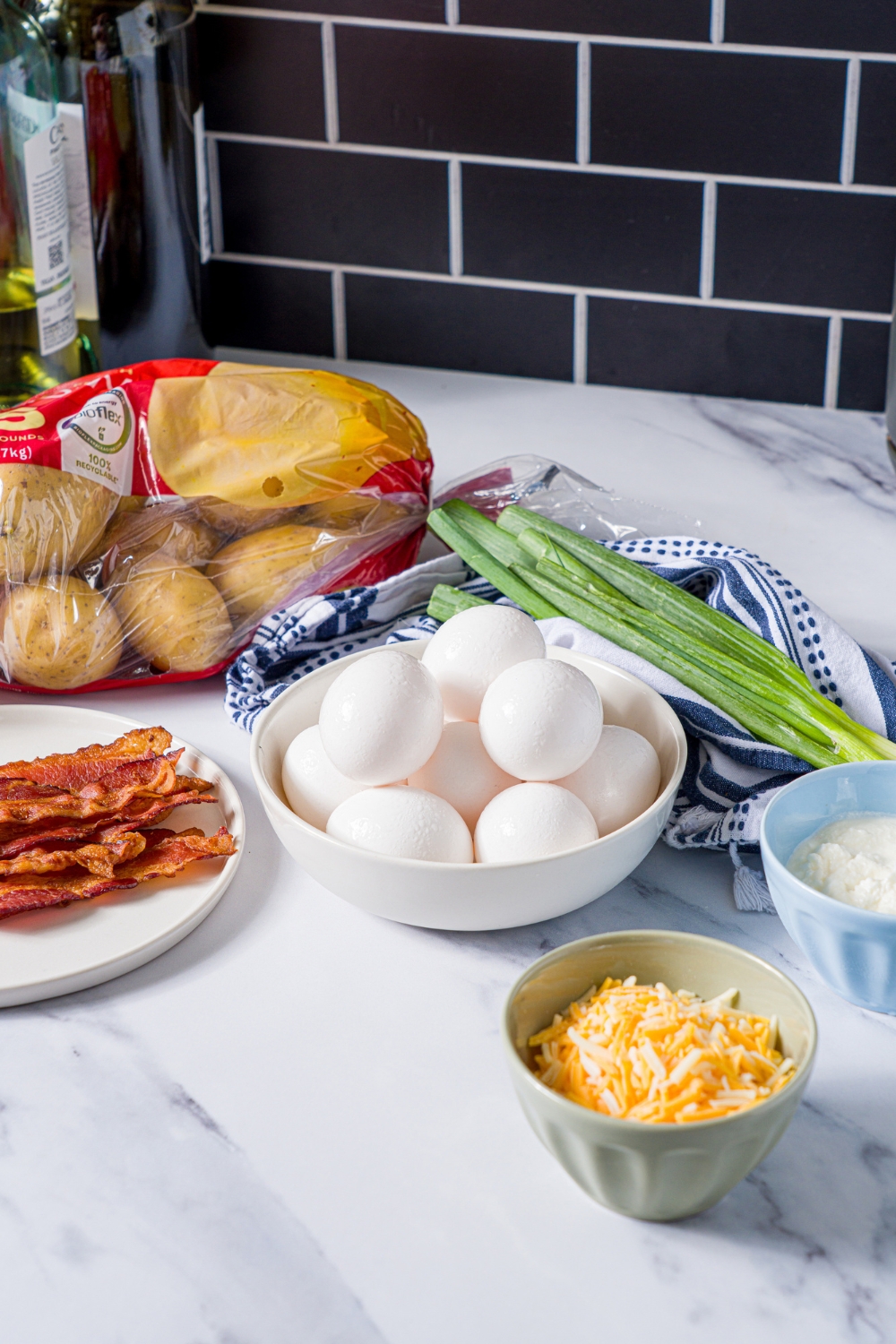 A marble counter with ingredients to make potato crust quiche including potatoes, eggs, bacon, shredded cheese, ricotta cheese, and green onion.