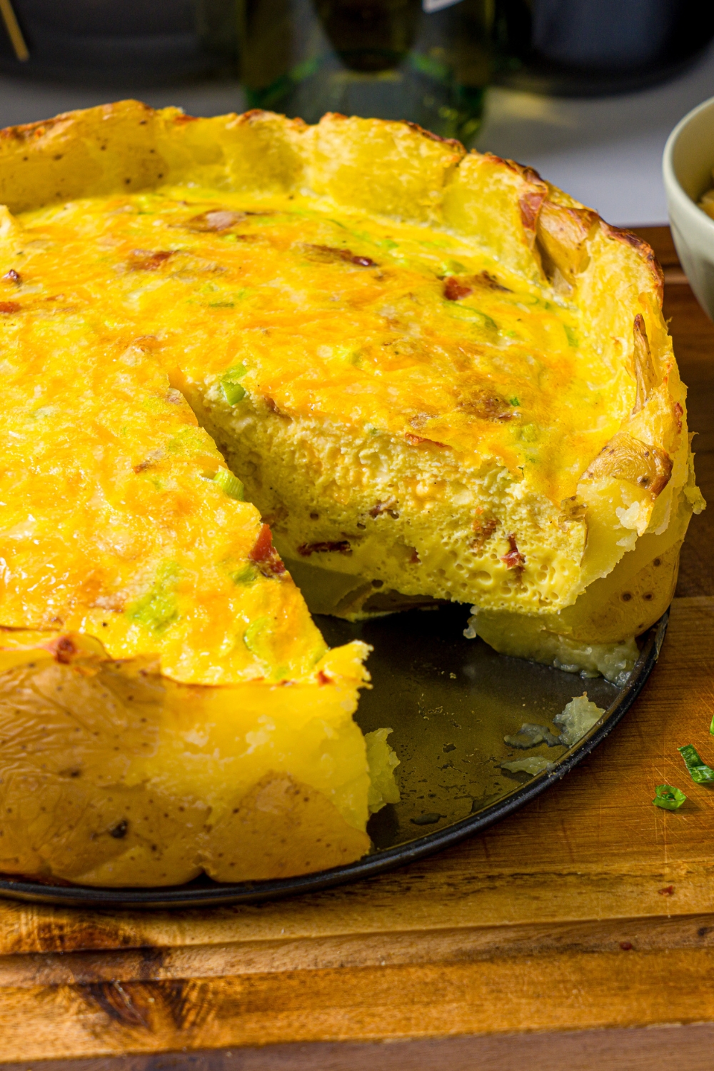 A wooden board with an open springform pan with baked potato crust quiche. A slice has been taken from the quiche.