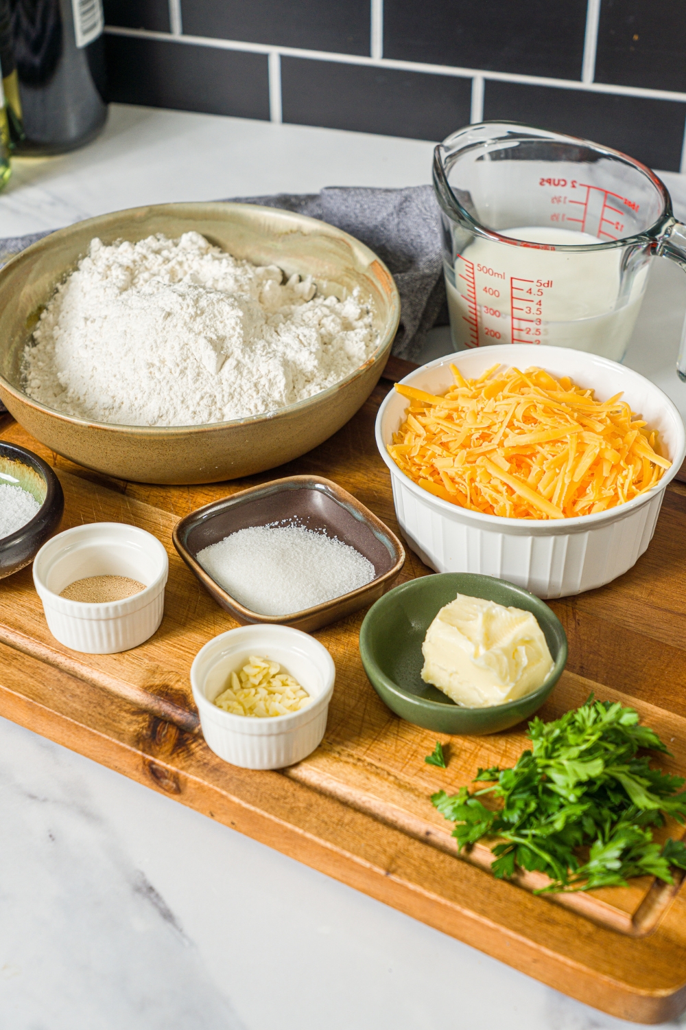 A wooden board with several bowls of ingredients to make pull apart cheesy garlic bread including flour, shredded cheddar cheese, butter, milk, yeast, and more.
