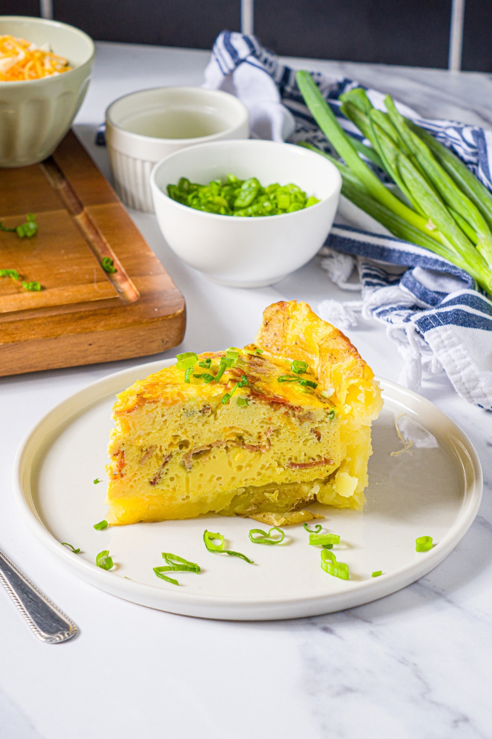 A white plate with a slice of potato crust quiche garnished with sliced green onion. The plate is on a marble counter with a fork and bowls of garnishes.