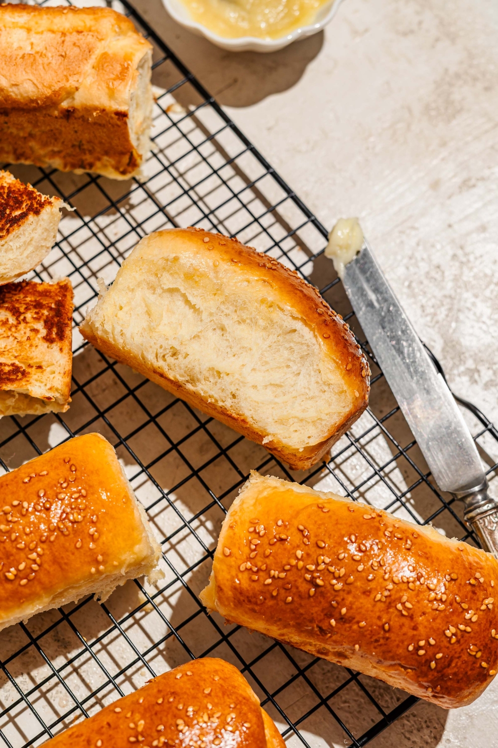 A wire rack with several pieces of baked Raising Cane's bread topped with sesame seeds. One of the loaves is on its side. The wire rack is on a tan counter with a buttered knife.
