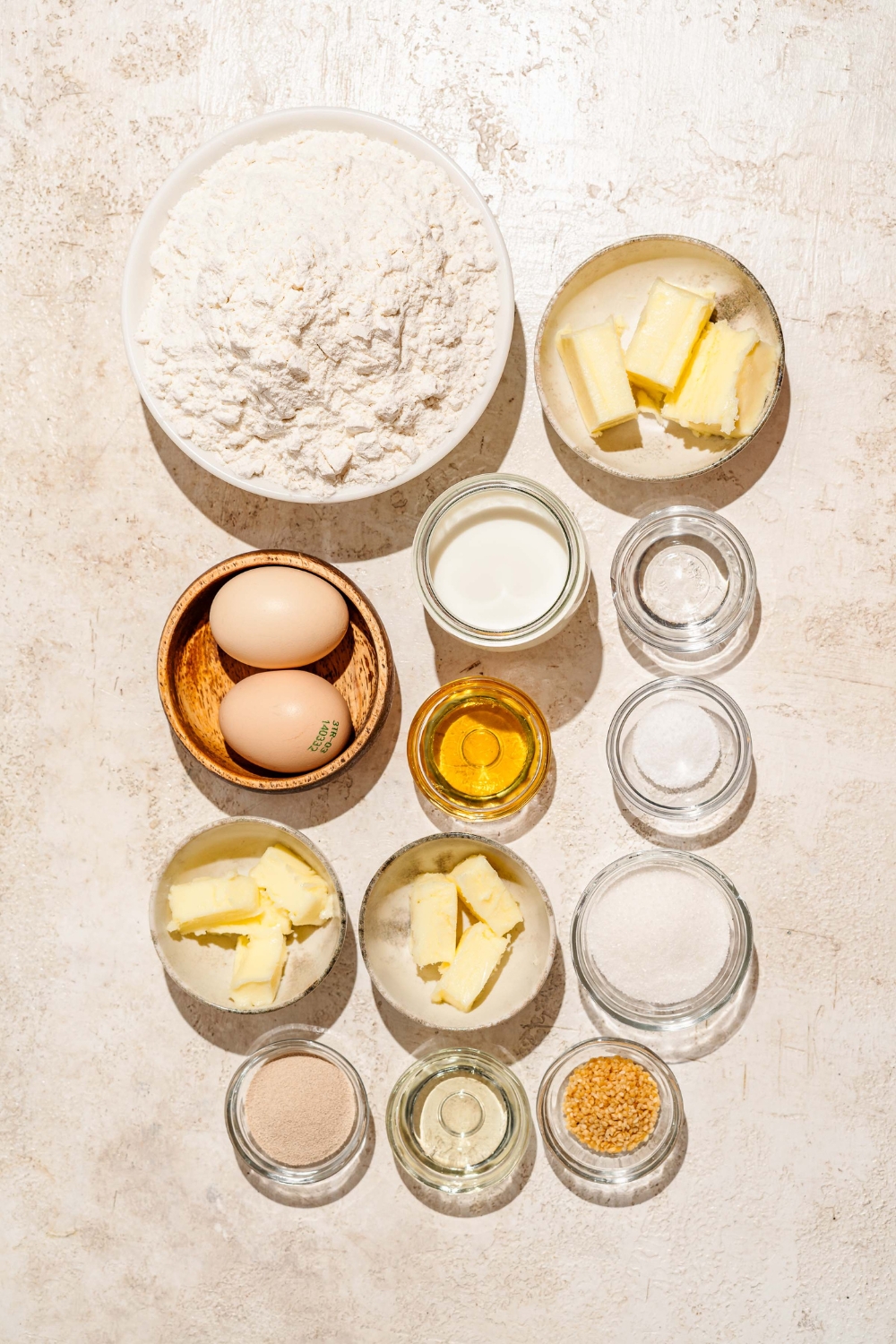 A tan counter with several bowls containing ingredients to make Raising Cane's bread including flour, butter, egg, sugar, yeast, milk, oil, water, butter, sesame seeds, and more.
