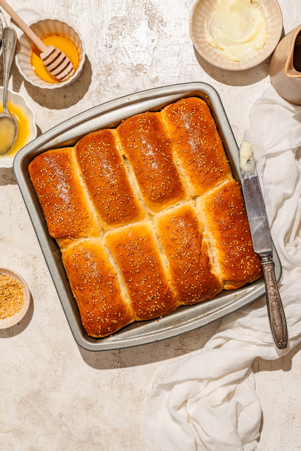 A large loaf pan with baked pieces of Raising Cane's bread topped with sesame seeds and finished with honey glaze. The pan is on a tan counter with a buttered knife and bowls of ingredients.