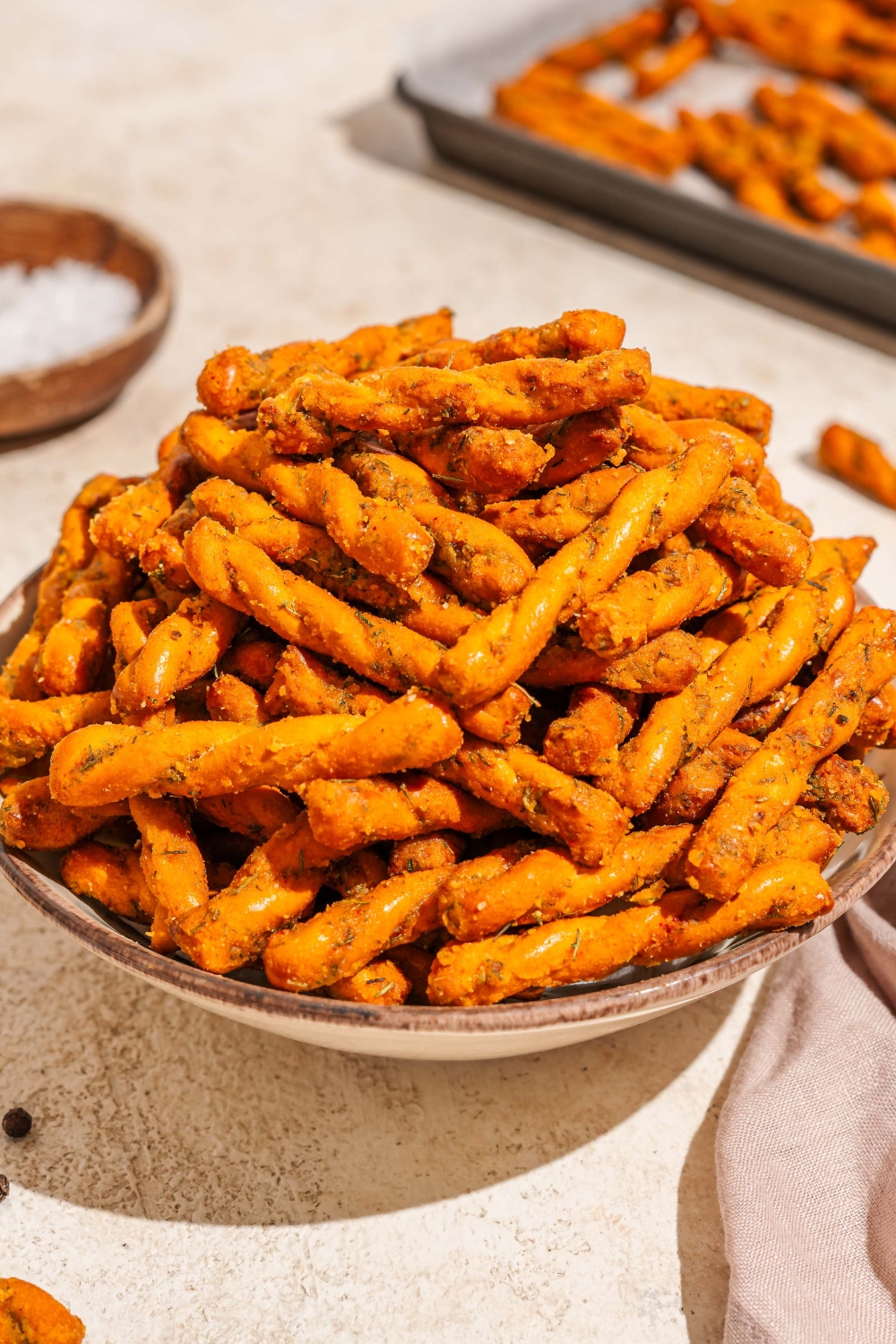 A bowl of seasoned pretzel braids. The bowl is on a tan counter with a white cloth napkin and baking sheet of pretzels.