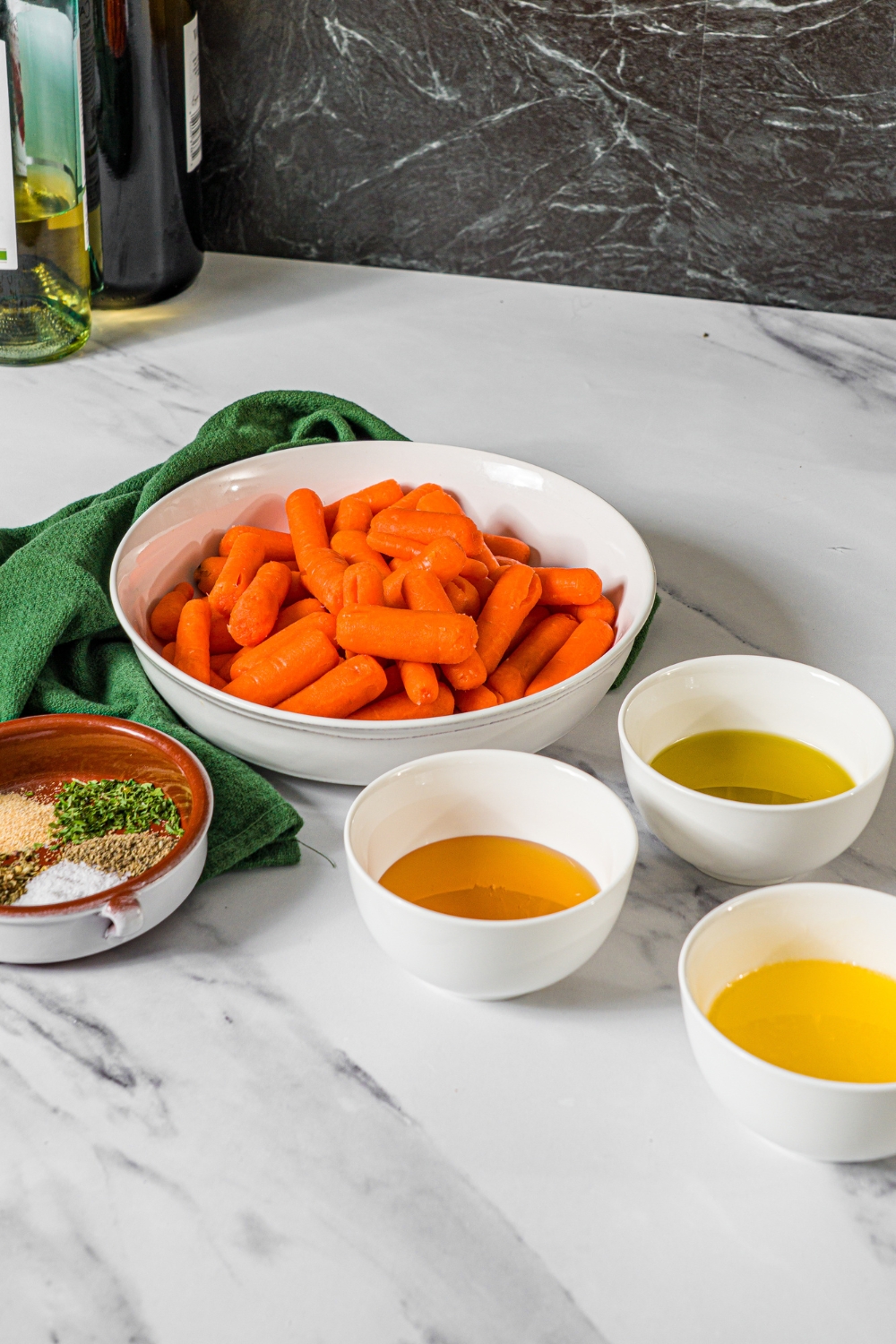 A marble counter with ingredients to make roasted baby carrots including baby carrots, honey, oil, butter, and seasonings.