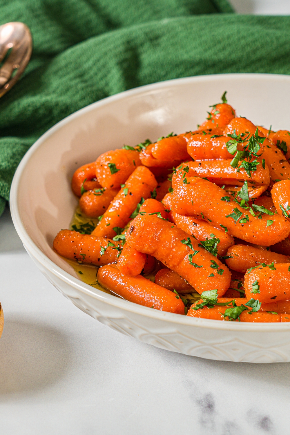 A bowl of honey roasted baby carrots finished with fresh parsley. The bowl is on a marble counter with a green cloth napkin.