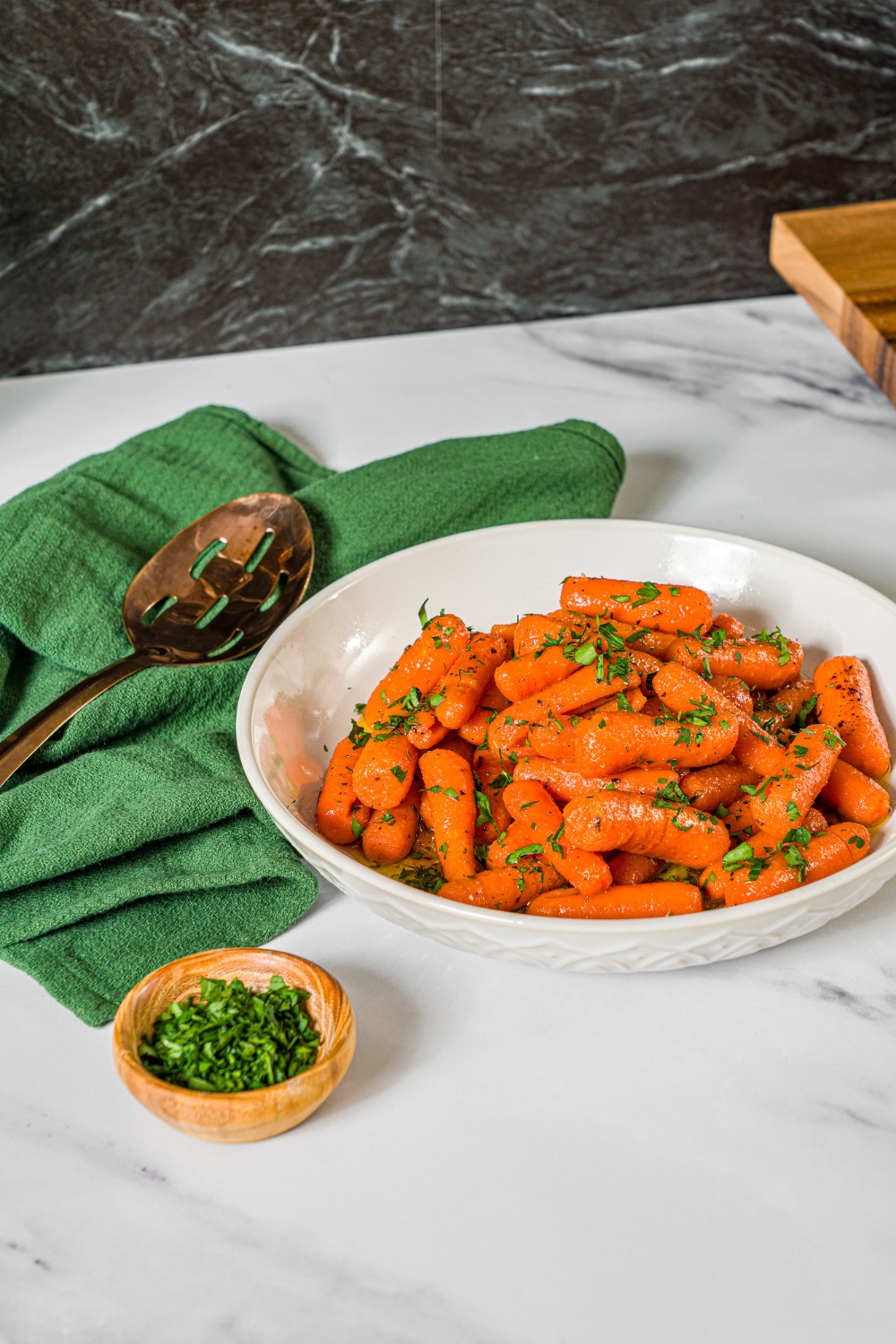 A white bowl with honey roasted baby carrots finished with fresh parsley. The bowl is on a marble counter with a small bowl of parsley and green cloth napkin.