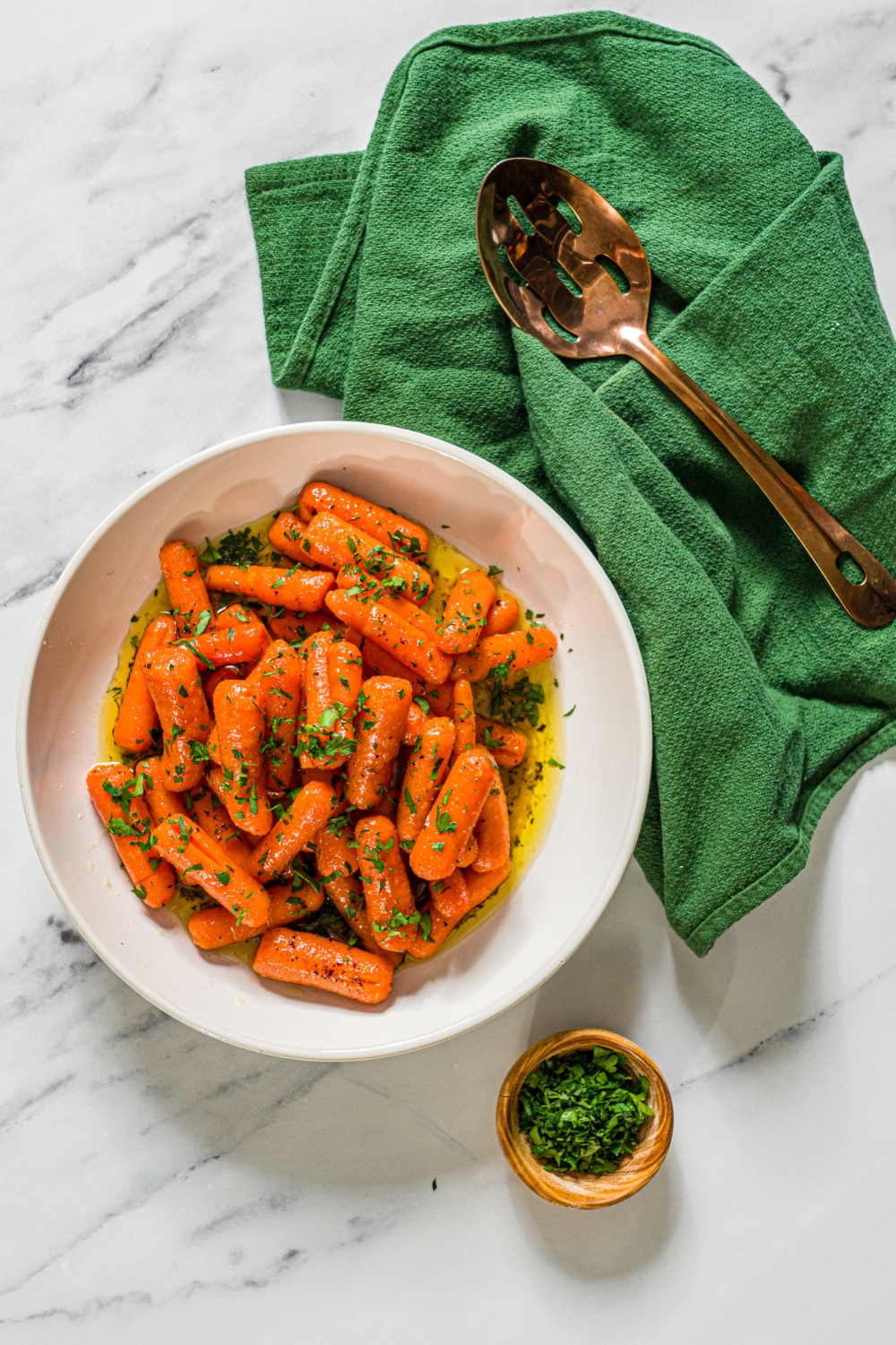 A white bowl with honey roasted baby carrots finished with fresh parsley. The bowl is on a marble counter with a small bowl of parsley and green cloth napkin.