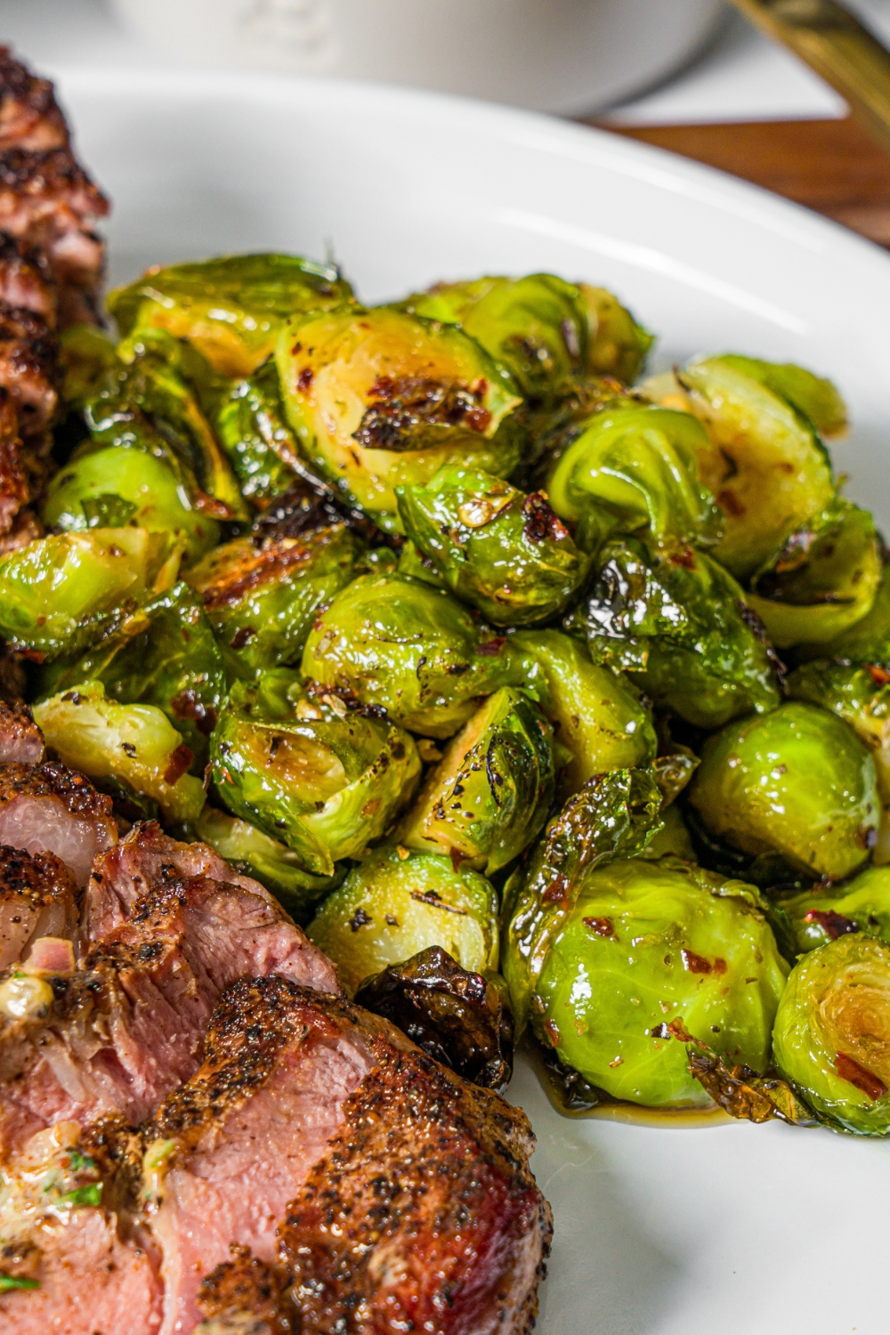 A white plate with roasted sweet chili brussels sprouts with sliced grilled steak.