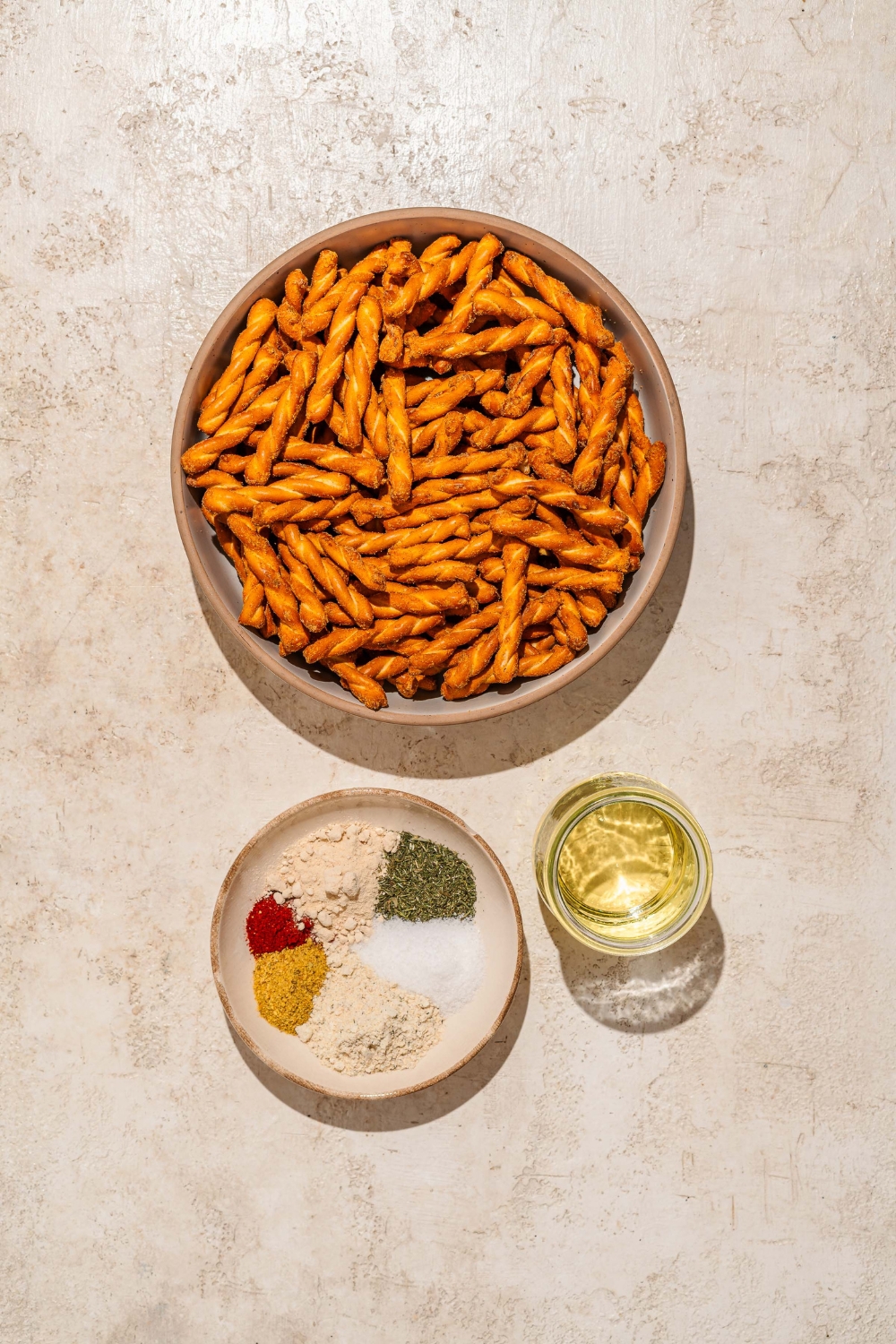 A tan counter with bowls containing ingredients to make seasoned pretzels including of pretzel braids, seasonings, and oil.