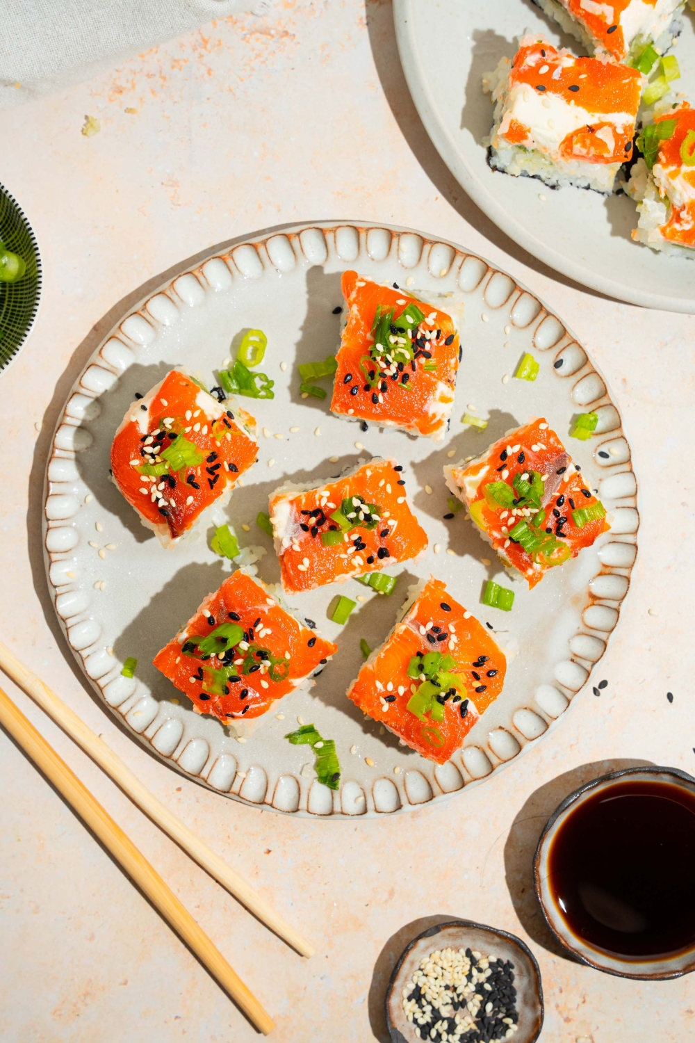 A plate of lazy sushi with salmon topped with sliced green onion and sesame seeds. The plate is on a tan counter with a plate of soy sauce and sesame seeds with an additional plate of sushi.
