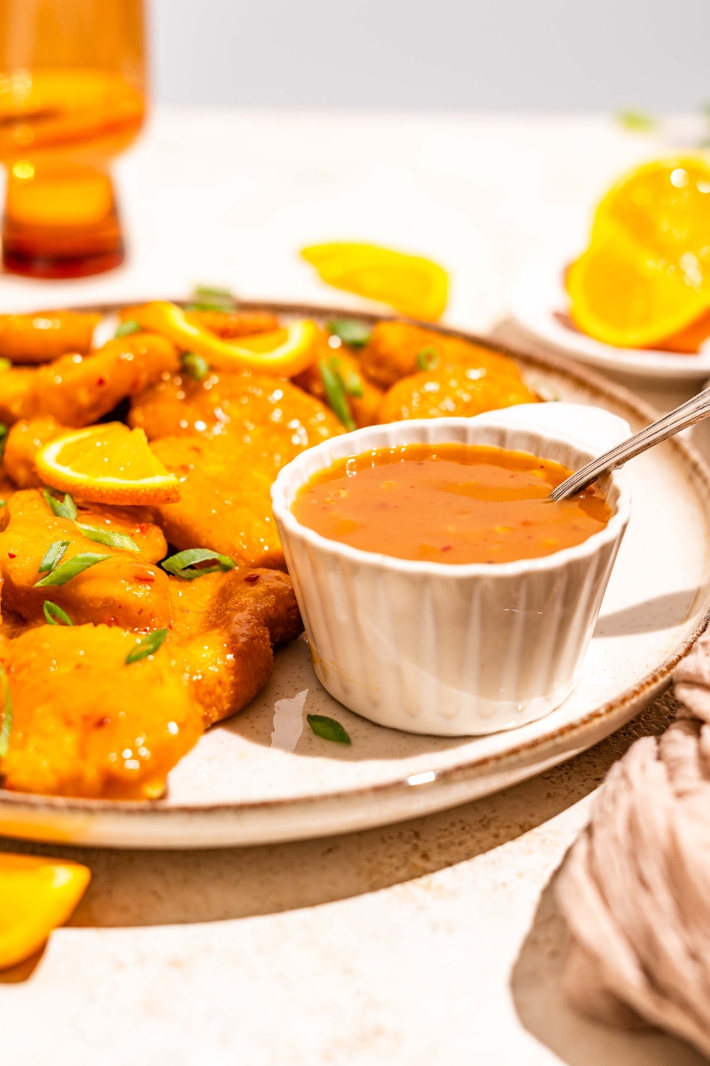 A white plate with chicken tossed in orange sauce and garnished with sliced green onion and orange slices. The plate is served with a small bowl of orange sauce with a spoon in the bowl. The plate is on a tan counter with a brown cloth napkin.