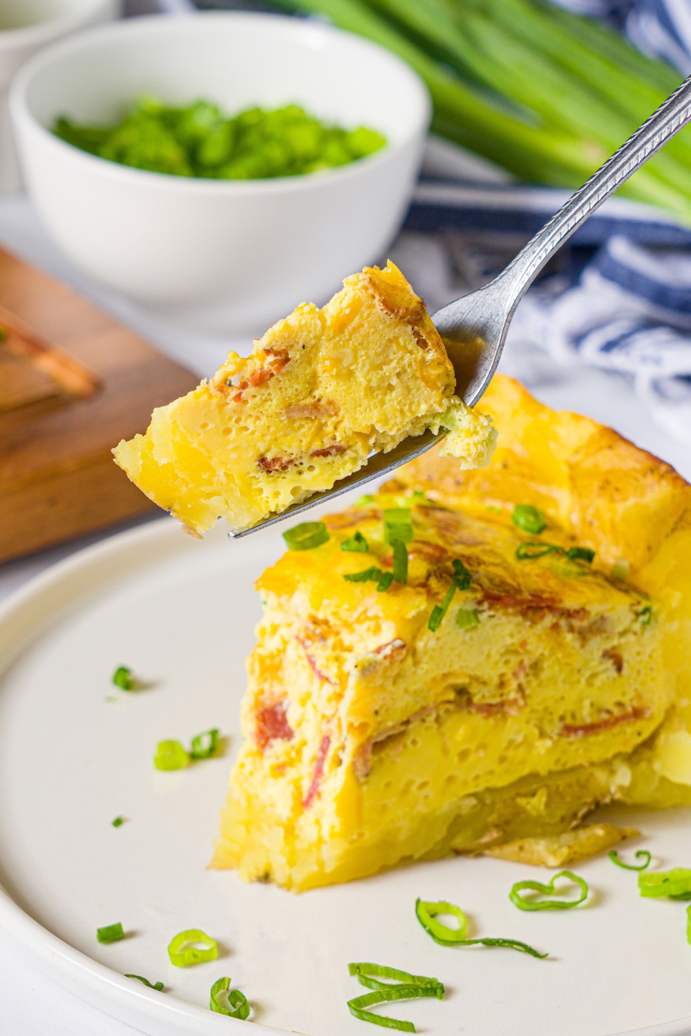 A white plate with a slice of potato crust quiche garnished with sliced green onion. A fork is taking a bite from the quiche. The plate is on a marble counter with a bowl of garnishes.
