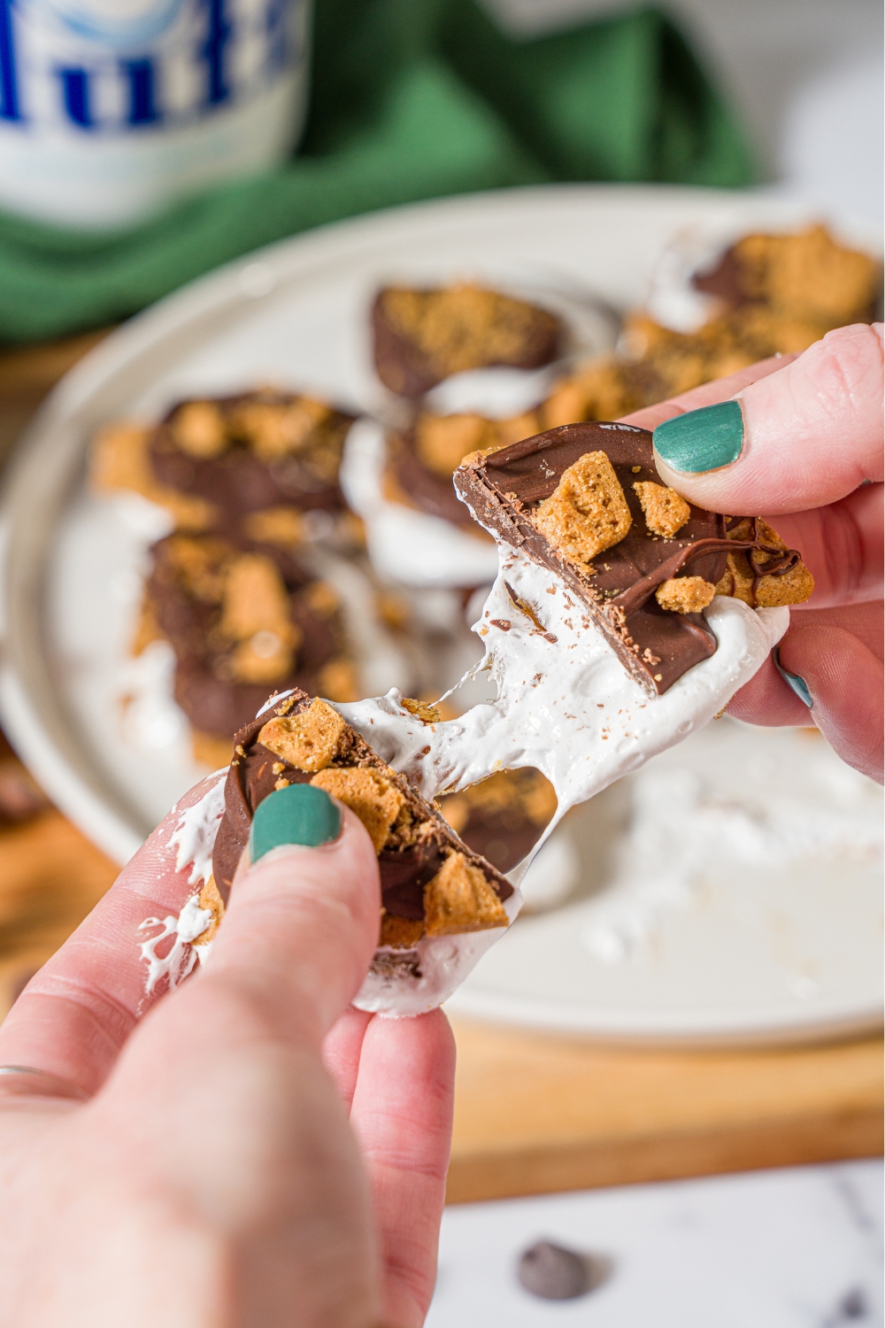 Two hands pulling apart, a smash date s'more. The hands are holding this above a white plate that is filled with the date s'mores.
