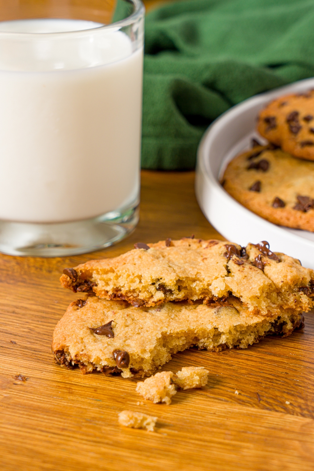 A chocolate cookie without baking soda broken in half. The cookie is on a wooden counter with a plate of cookies and a glass of milk.