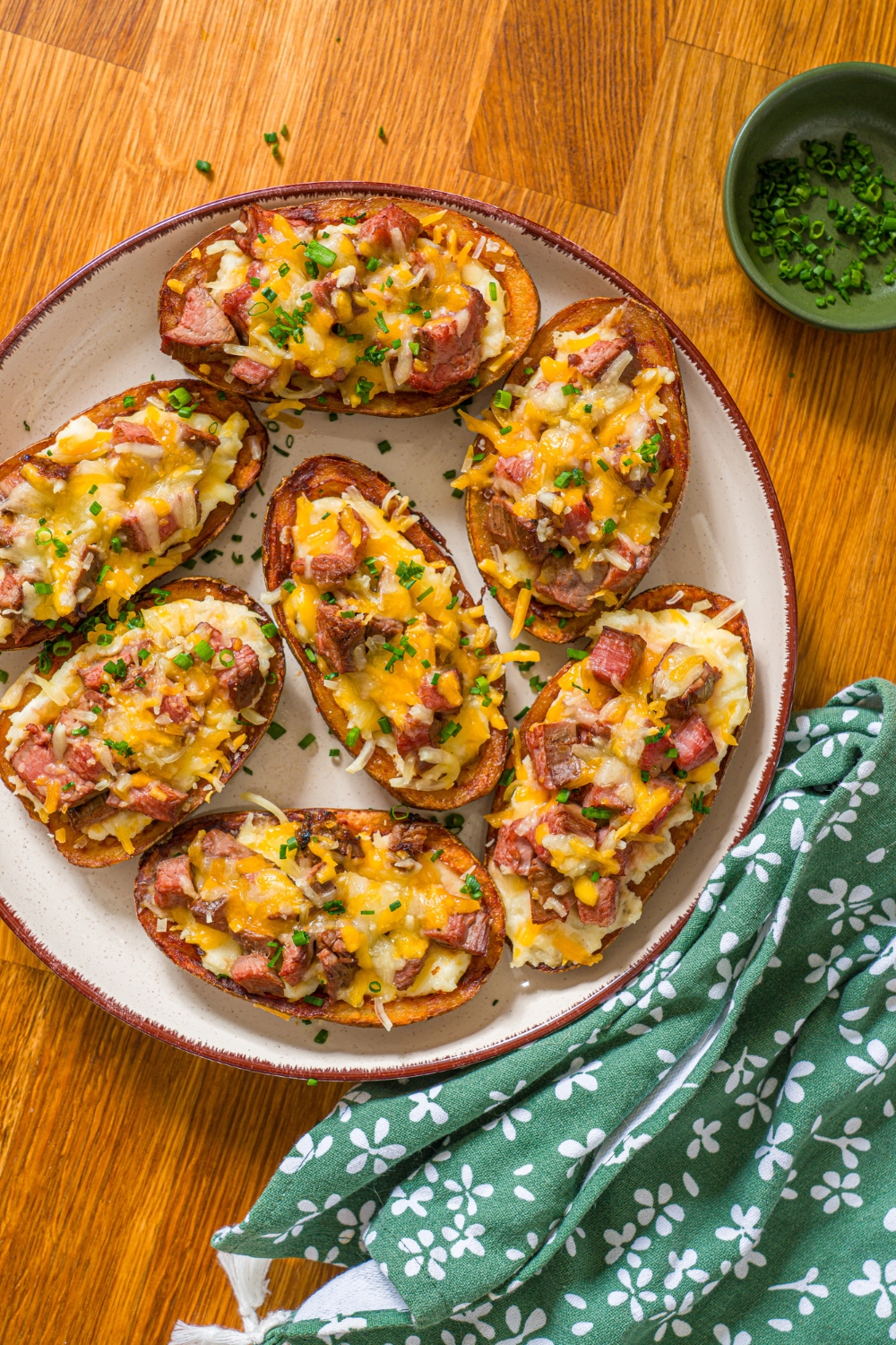 A white plate with several steak loaded potato skins with melted cheese on top and garnished with sliced chives. The plate is on a wooden counter with a green floral napkin.