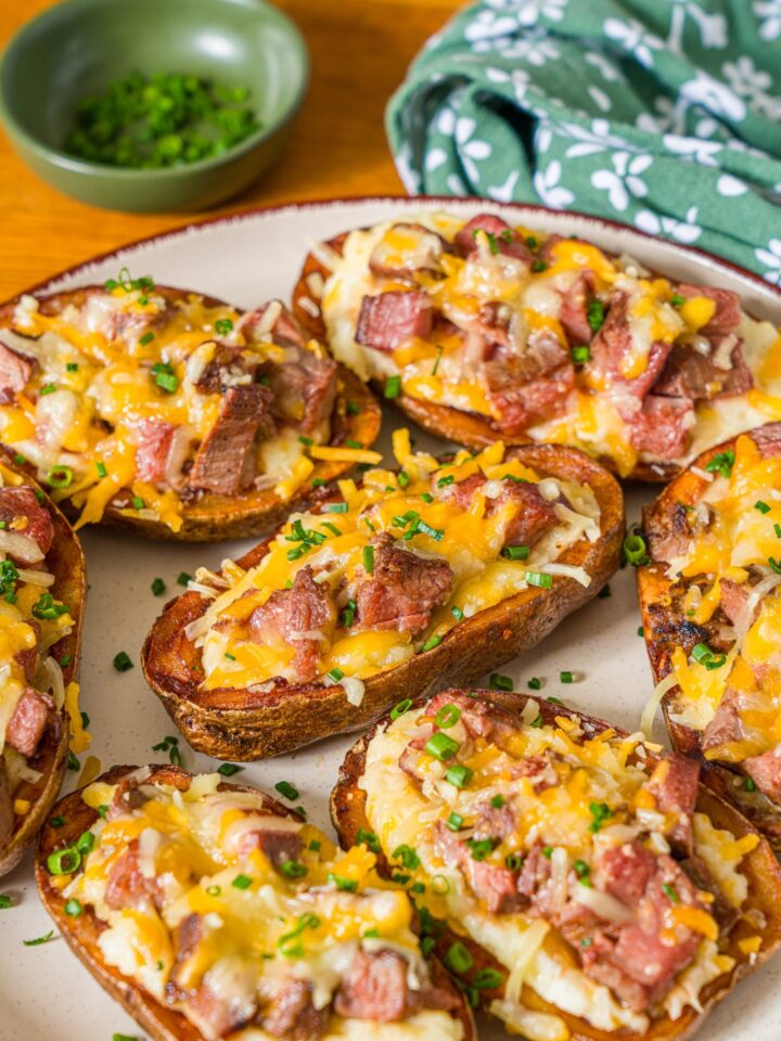 A white plate with several steak loaded potato skins with melted cheese on top and garnished with sliced chives. The plate is on a wooden counter with a green floral napkin.