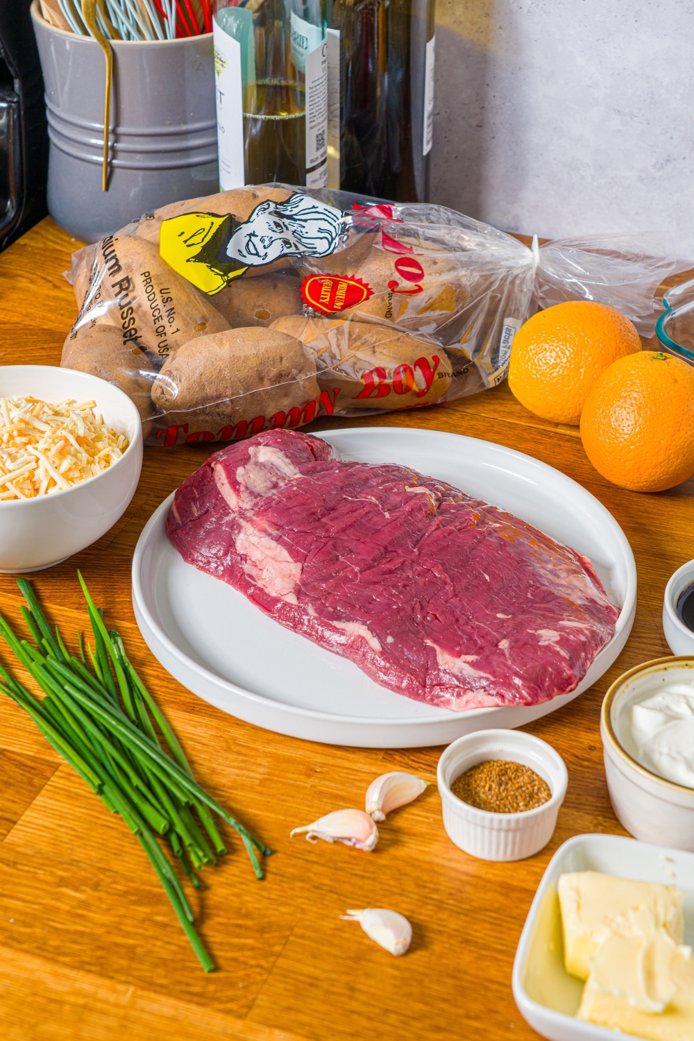 A wooden counter with ingredients to make steak loaded potato skins including steak, potatoes, butter, cheese, sour cream, and more.