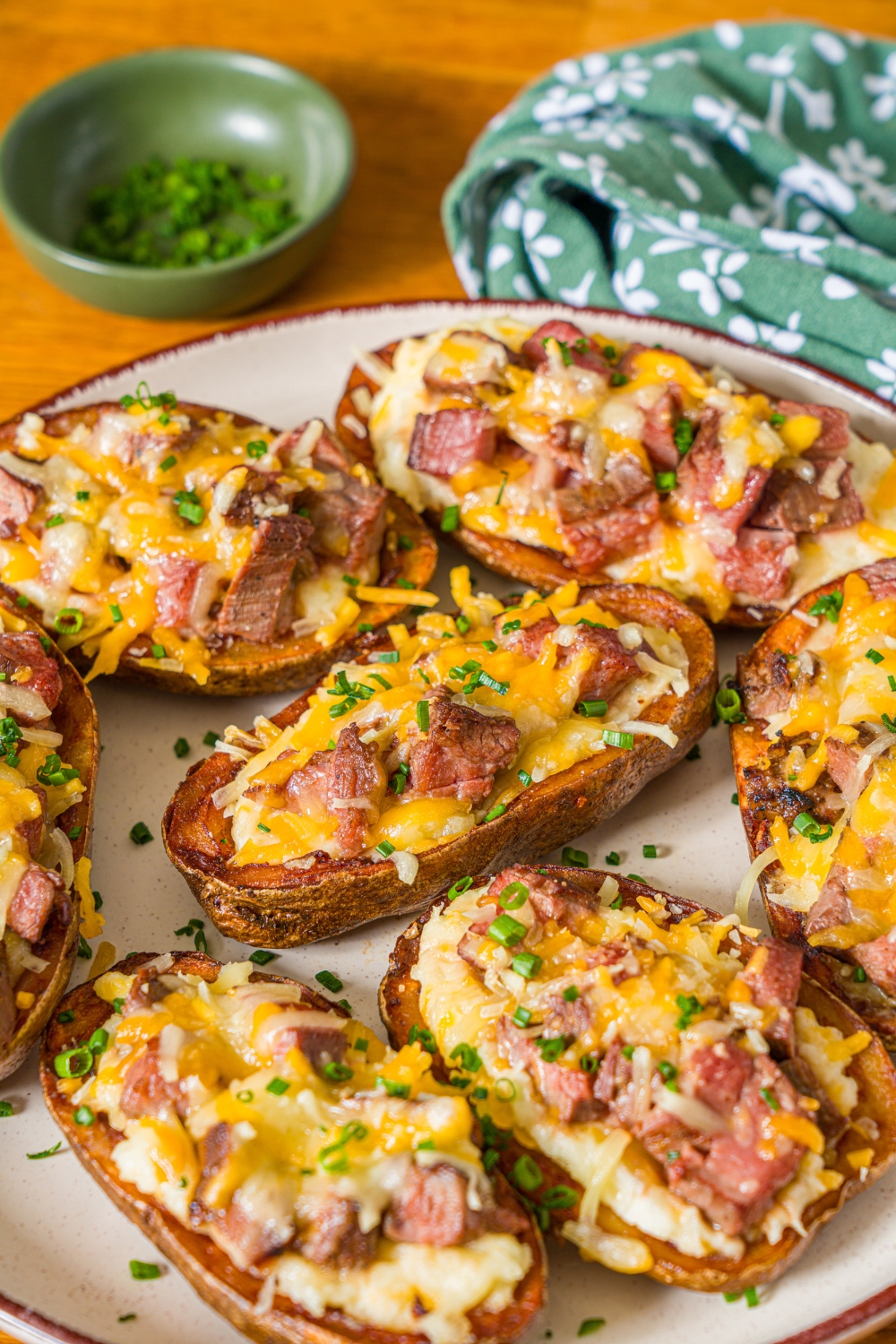 A white plate with several steak loaded potato skins with melted cheese on top and garnished with sliced chives. The plate is on a wooden counter with a green floral napkin.