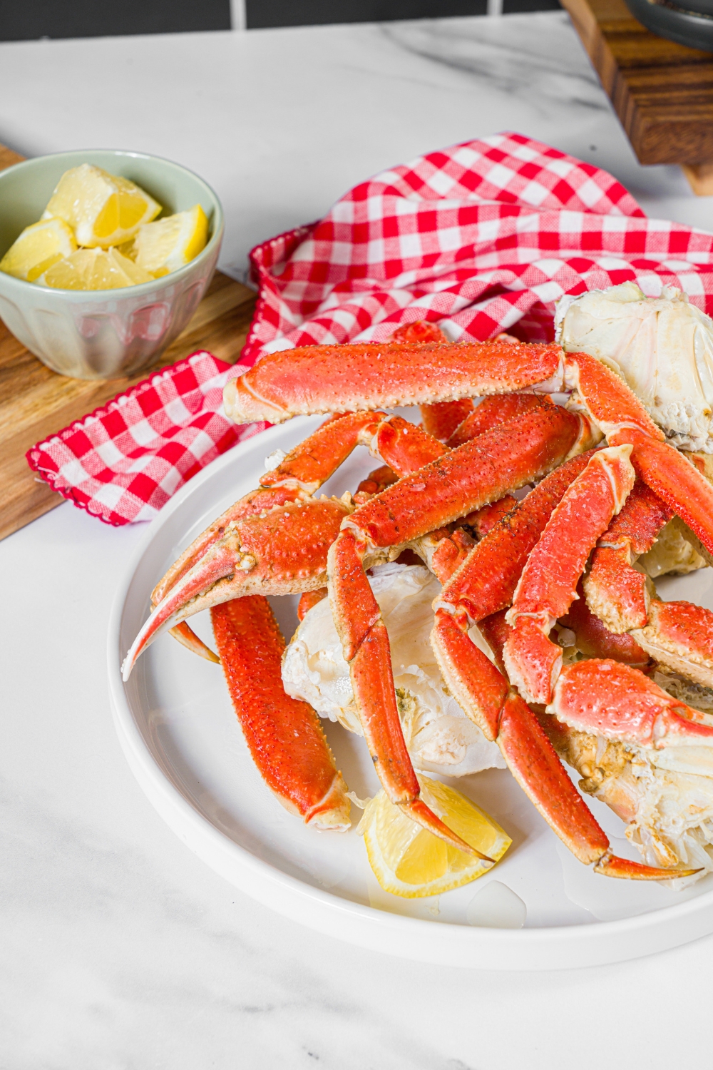 A white plate with steamed snow crab legs served with lemon. The plate is on a marble counter with a board of lemon butter sauce and lemons.
