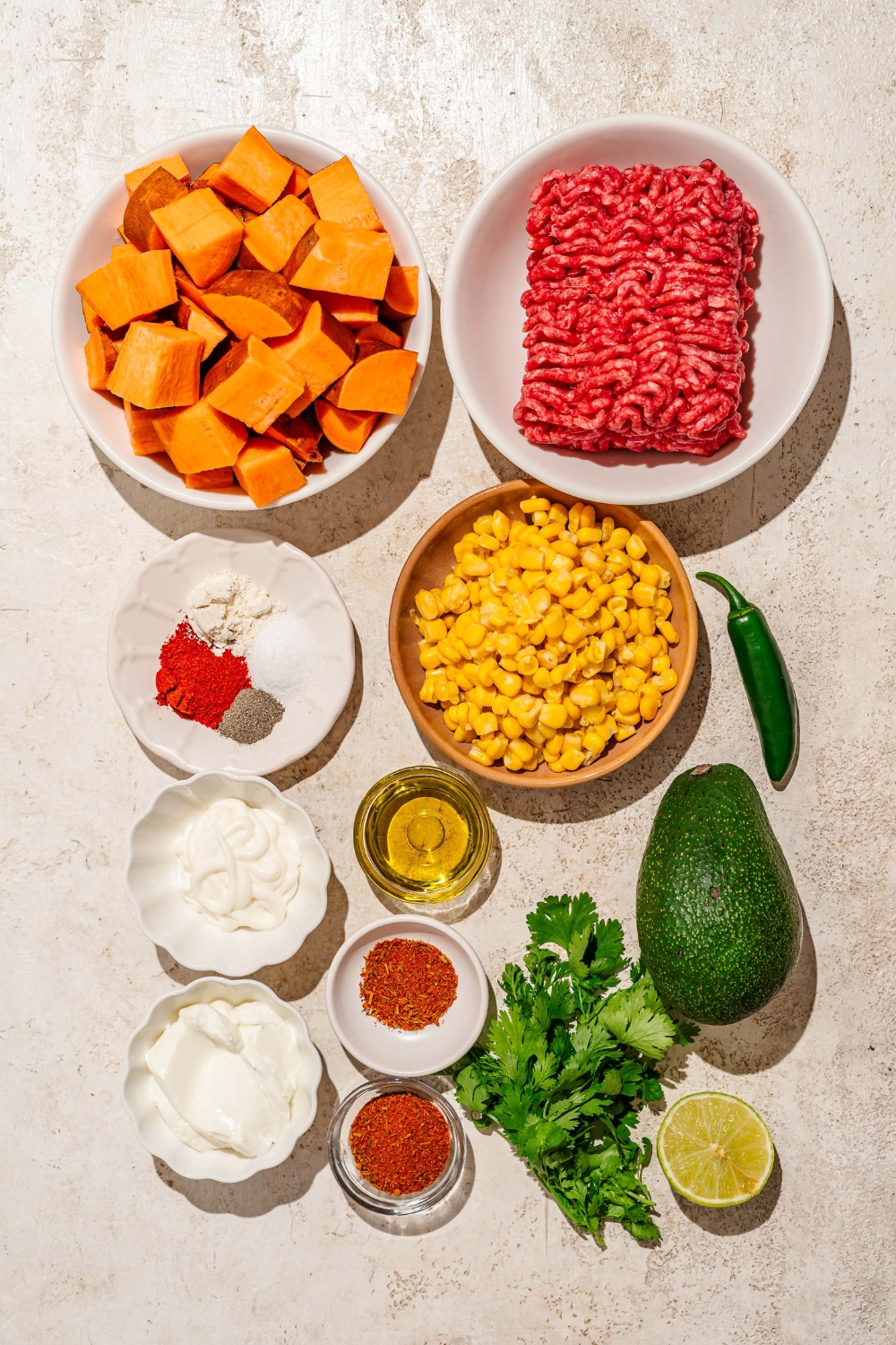 A tan counter with various bowls containing ingredients to make street corn beef bowl including street, ground beef, sweet potatoes, avocado, cilantro, lime, mayo, sour cream, and seasonings.