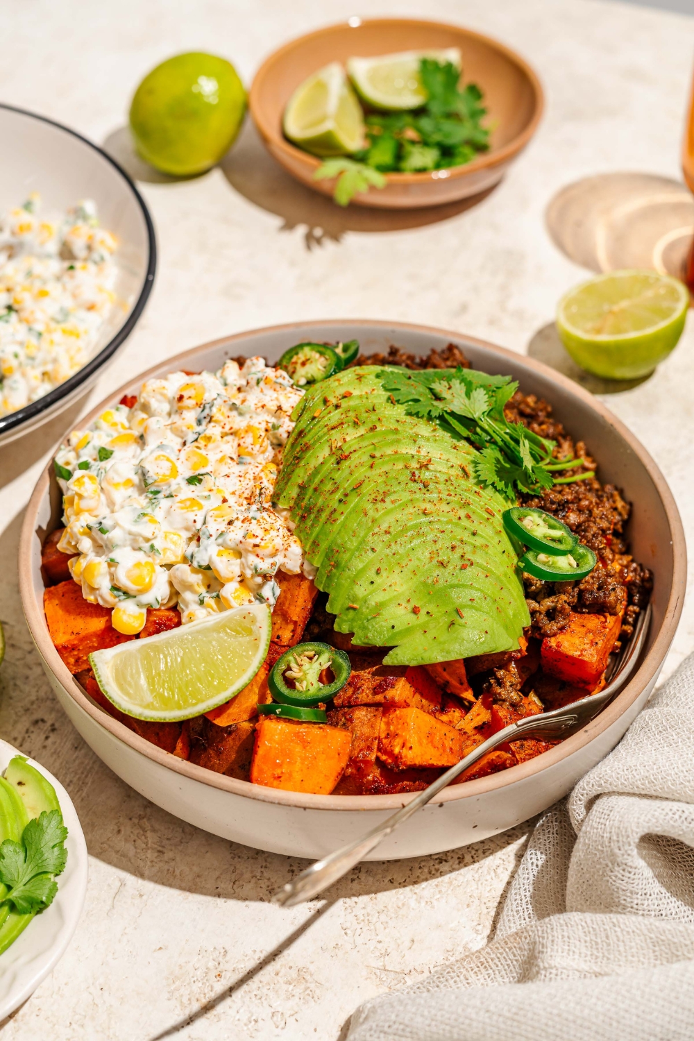 A bowl of street corn beef bowl including sweet potatoes, ground beef, street corn mixture, sliced avocado, jalapeños, and chile lime seasoning garnished with lime and cilantro. There is a fork in the bowl. The bowl is on a tan counter with a bowl of garnishes.