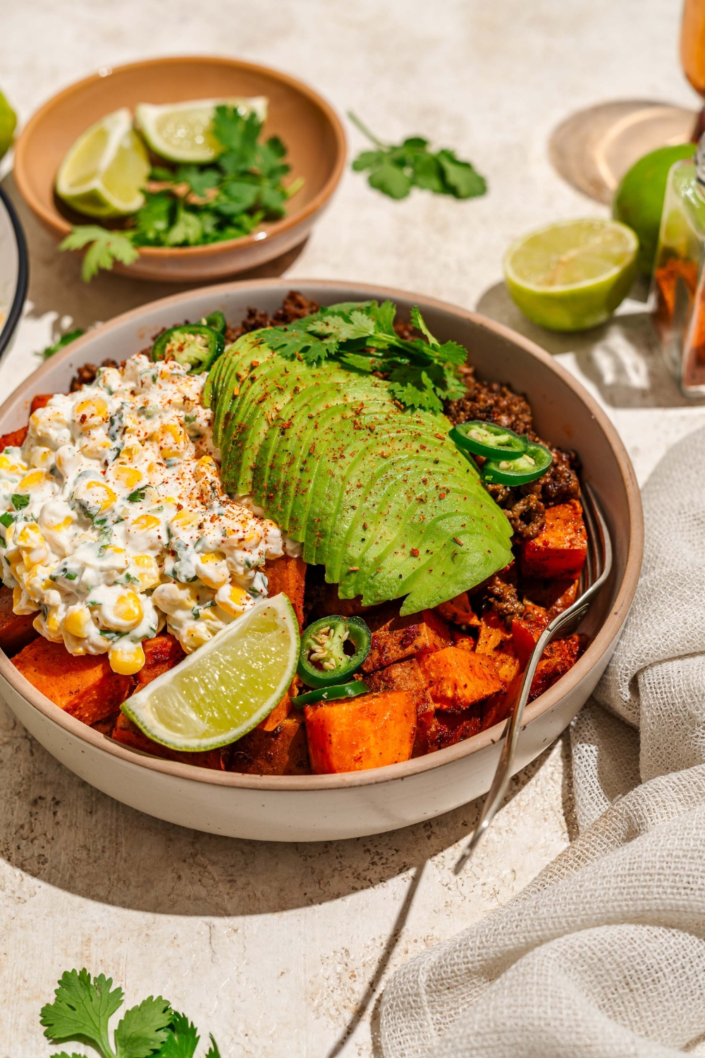 A bowl of street corn beef bowl including sweet potatoes, ground beef, street corn mixture, sliced avocado, jalapeños, and chile lime seasoning garnished with lime and cilantro. There is a fork in the bowl. The bowl is on a tan counter with a bowl of garnishes.