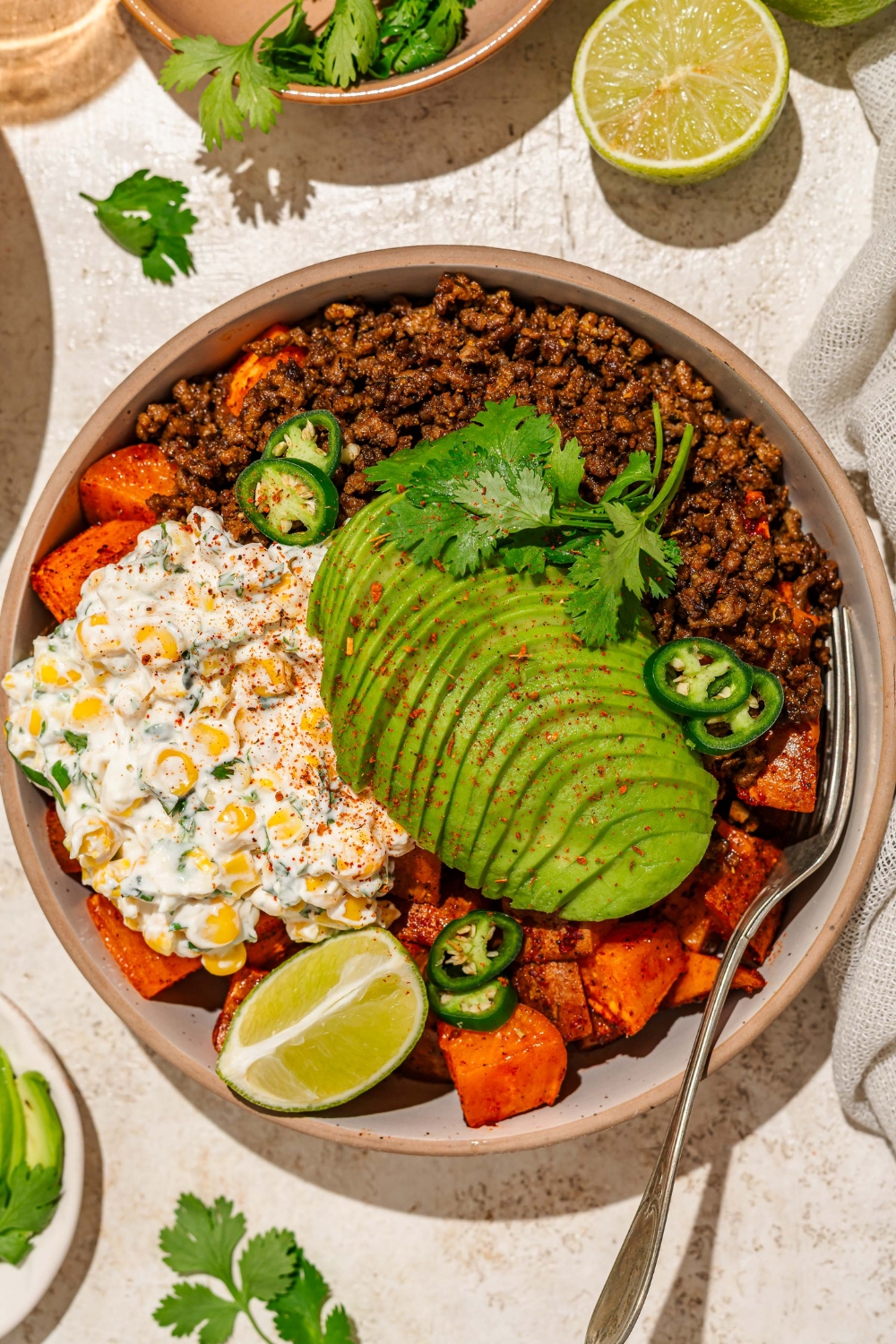 A bowl of street corn beef bowl including sweet potatoes, ground beef, street corn mixture, sliced avocado, jalapeños, and chile lime seasoning garnished with lime and cilantro. There is a fork in the bowl. The bowl is on a tan counter with a bowl of garnishes.