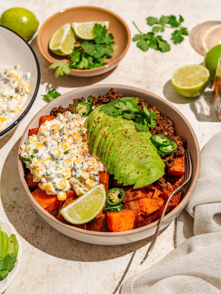 A bowl of street corn beef bowl including sweet potatoes, ground beef, street corn mixture, sliced avocado, jalapeños, and chile lime seasoning garnished with lime and cilantro. There is a fork in the bowl. The bowl is on a tan counter with a bowl of garnishes.