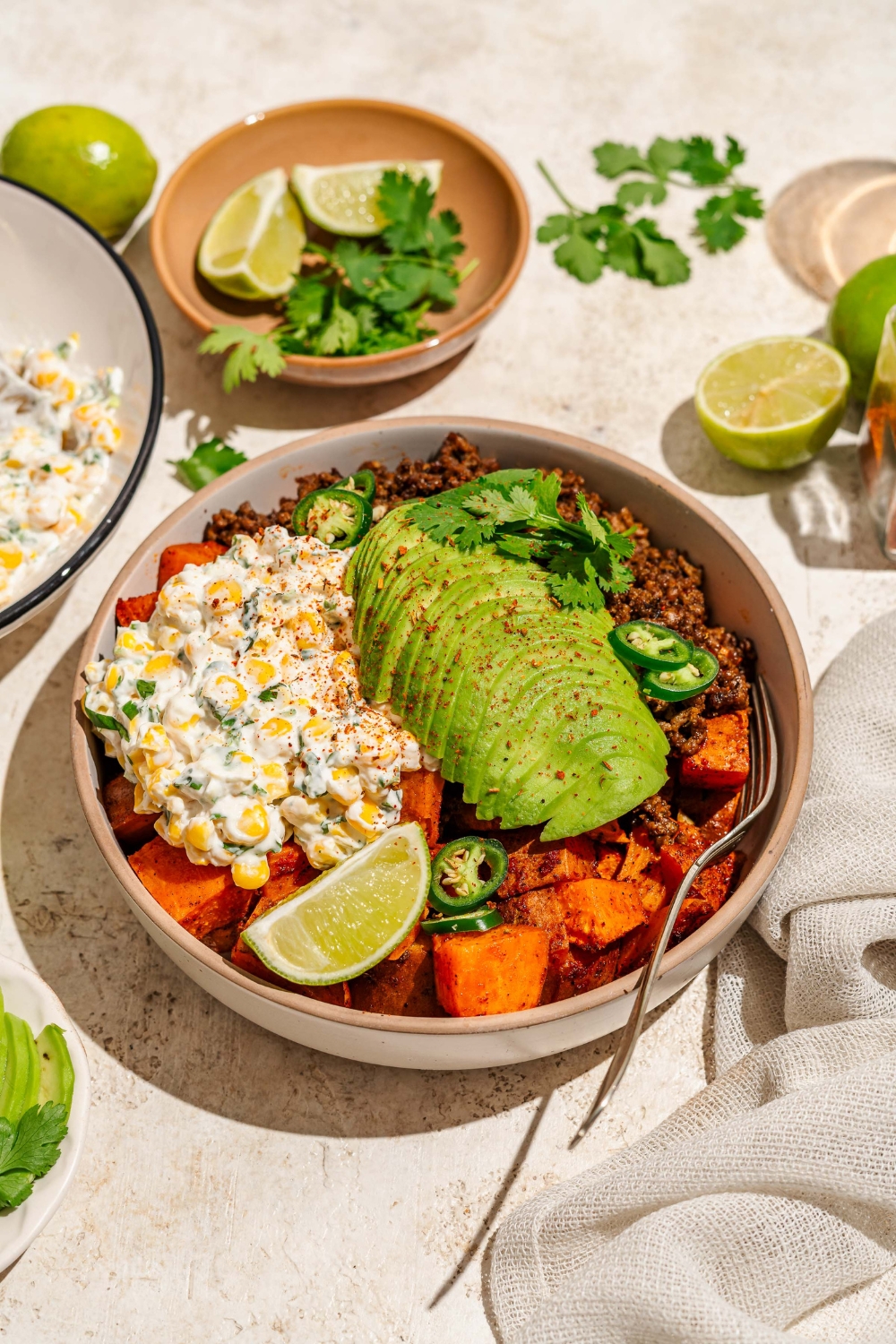 A bowl of street corn beef bowl including sweet potatoes, ground beef, street corn mixture, sliced avocado, jalapeños, and chile lime seasoning garnished with lime and cilantro. There is a fork in the bowl. The bowl is on a tan counter with a bowl of garnishes.