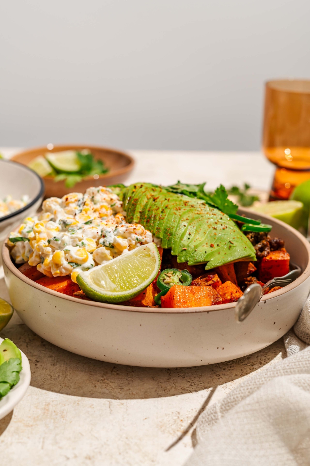 A bowl of street corn beef bowl including sweet potatoes, ground beef, street corn mixture, sliced avocado, jalapeños, and chile lime seasoning garnished with lime and cilantro. There is a fork in the bowl. The bowl is on a tan counter with a bowl of garnishes.