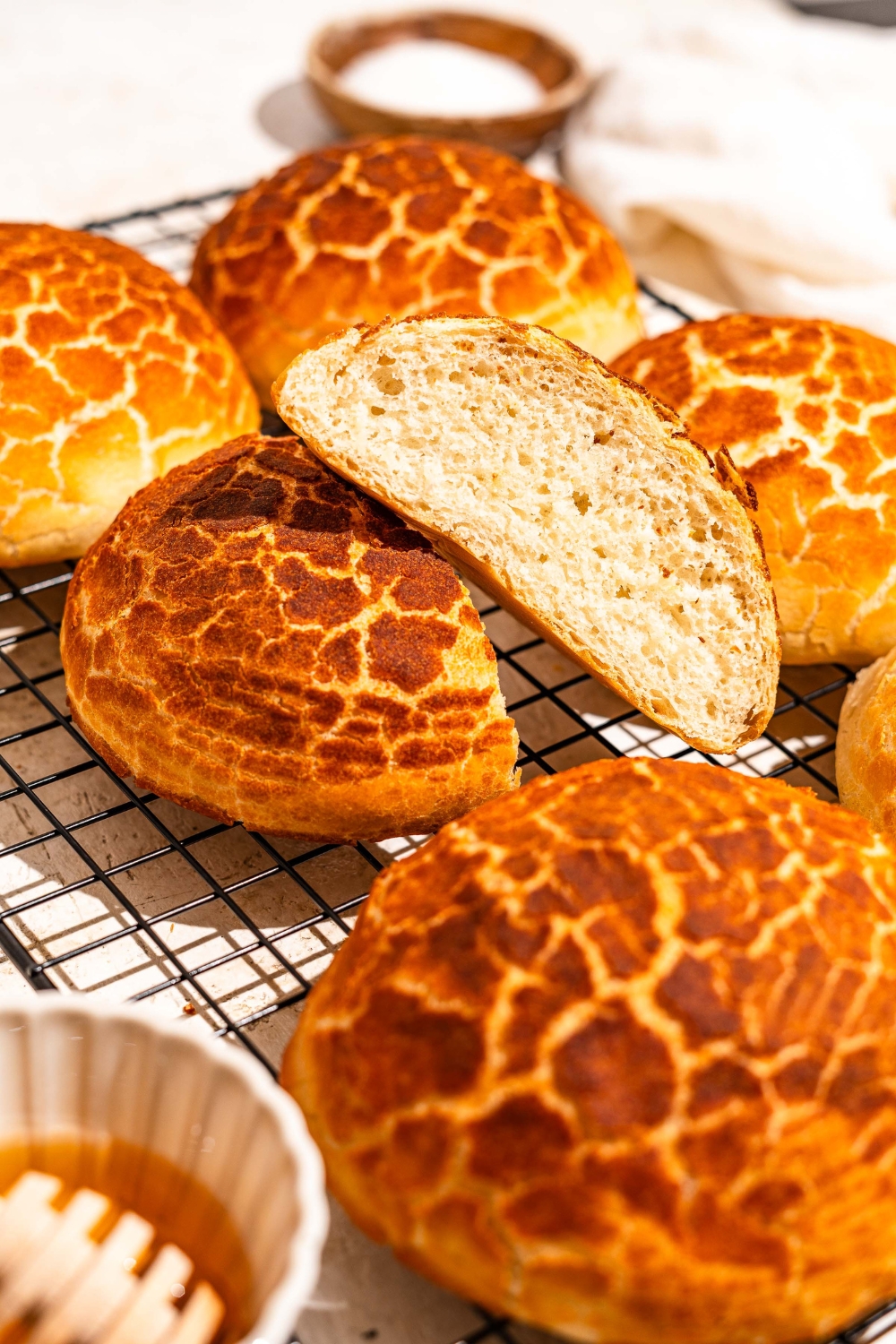A wire rack with baked Dutch Crunch bread loaves. One of the loaves is sliced in half. The rack is on a tan counter with a bowl of honey.