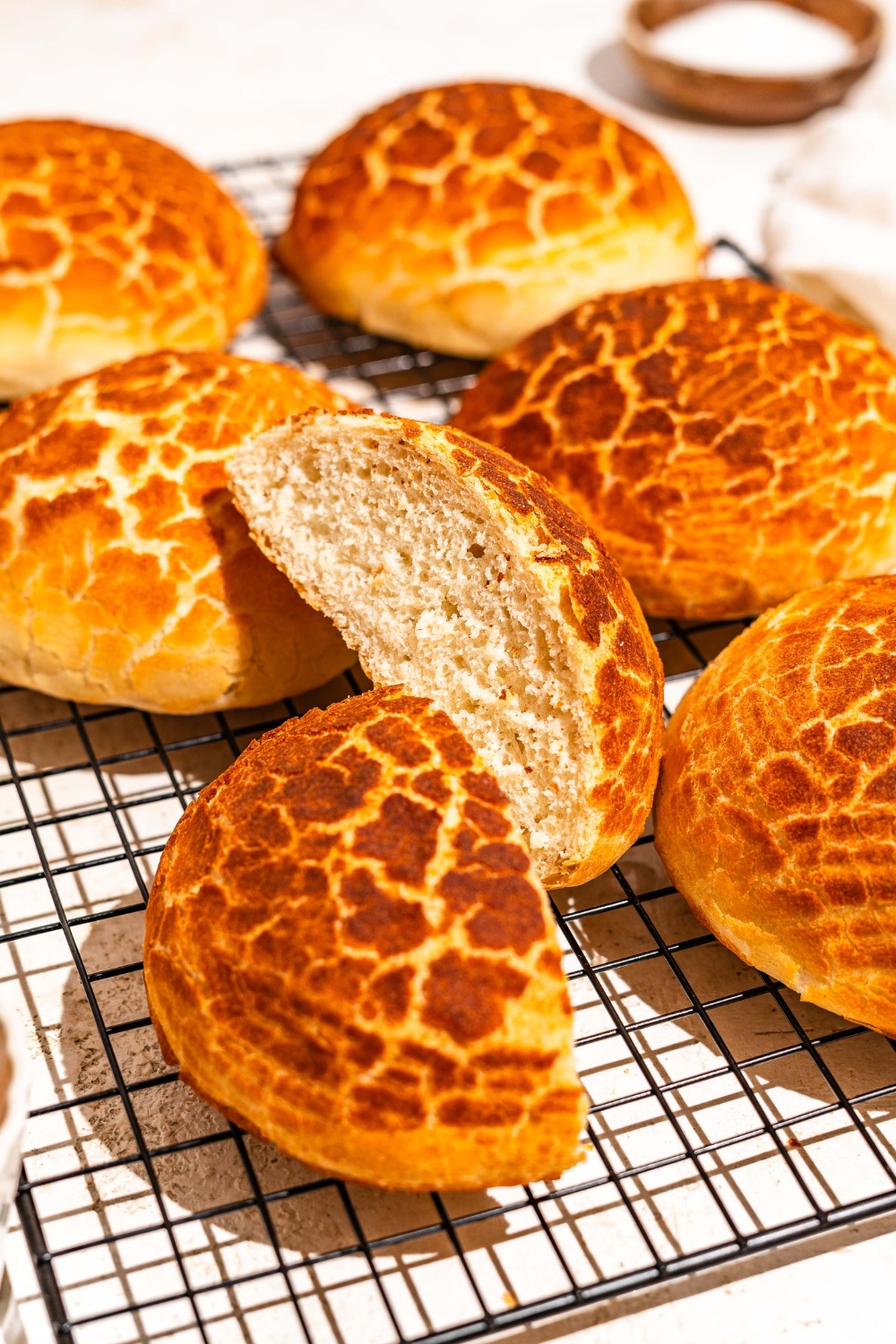 A wire rack with baked Dutch Crunch bread loaves. One of the loaves is sliced in half. The rack is on a tan counter with a bowl of honey.